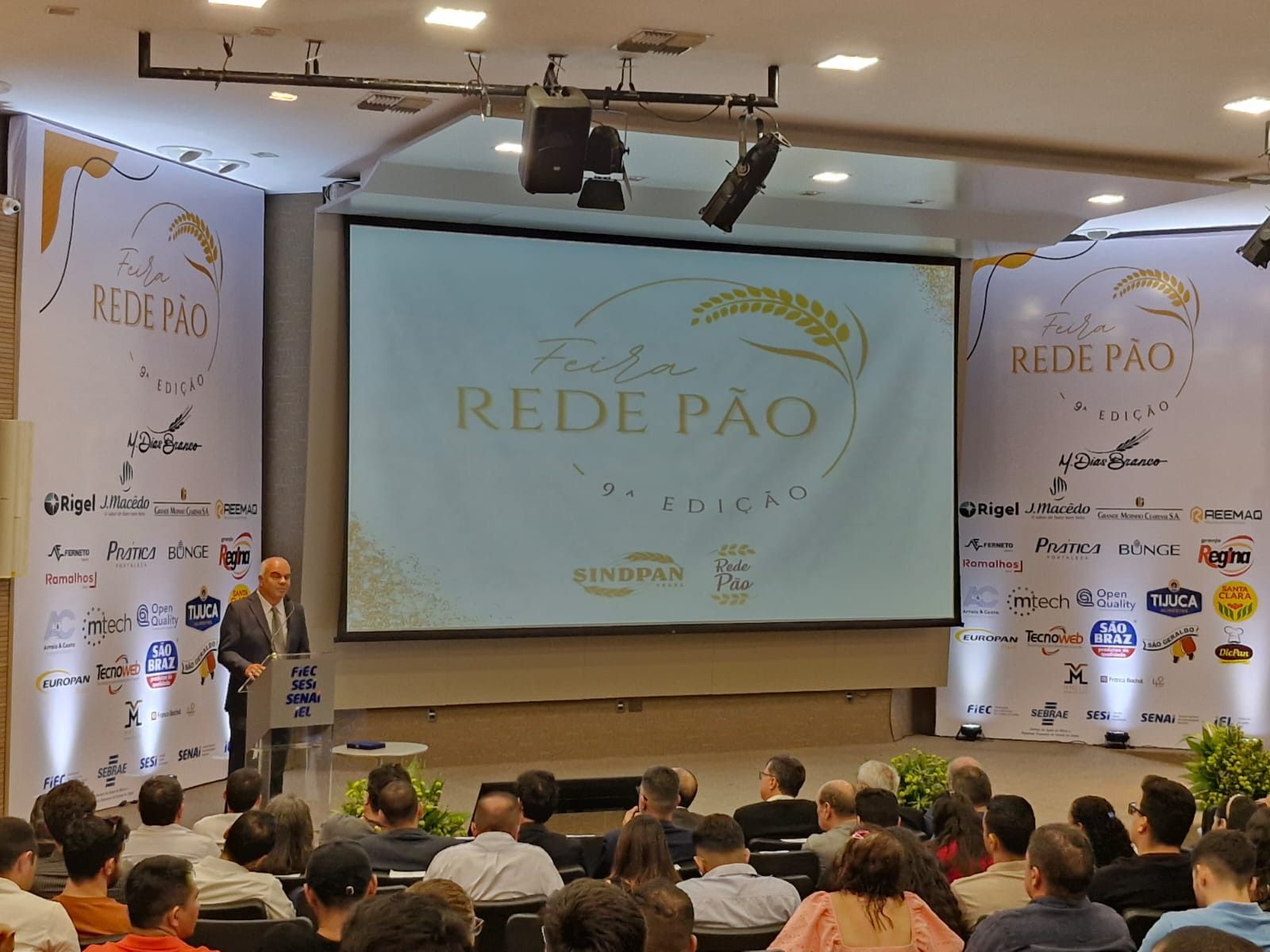 Um grupo de pessoas está sentado em frente a uma tela grande que diz rede pao