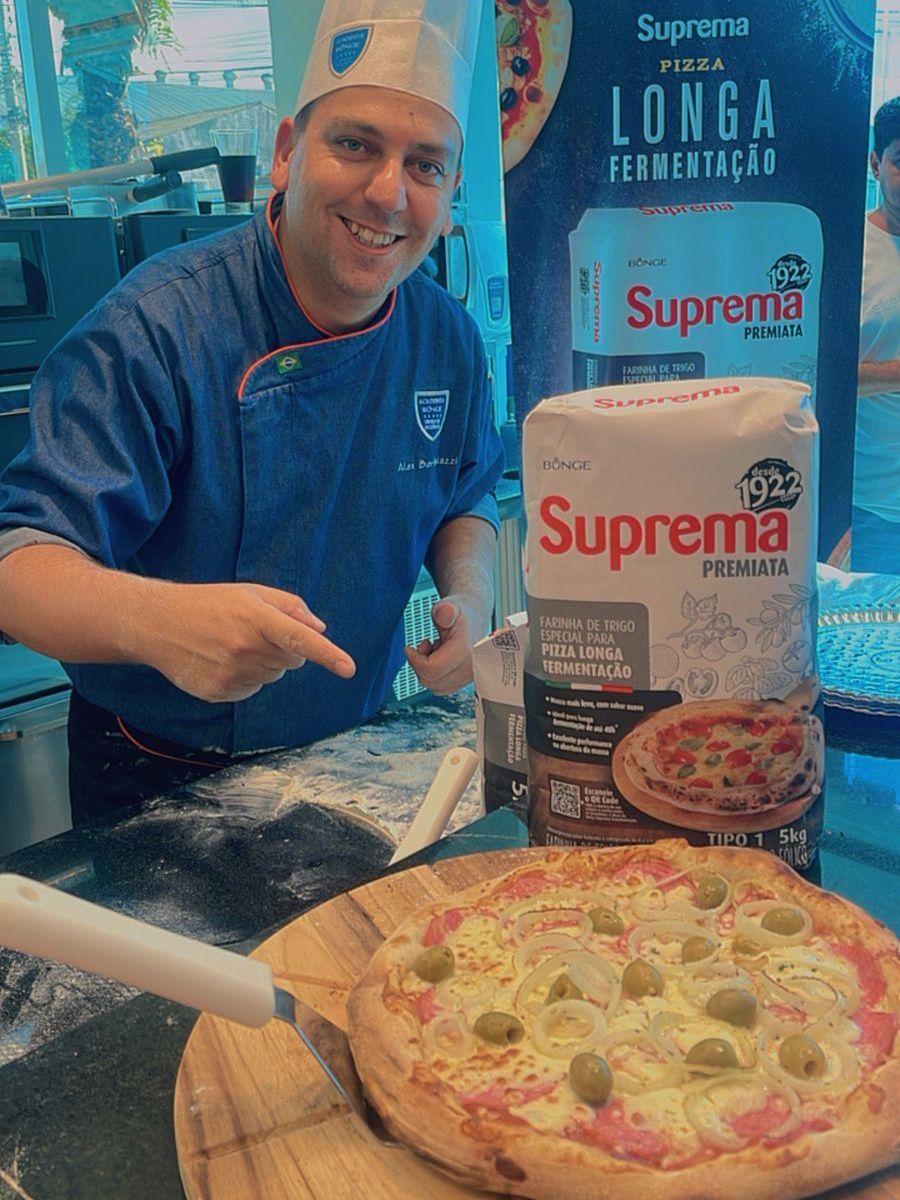 Um homem está de pé ao lado de uma pizza em uma tábua de corte de madeira.