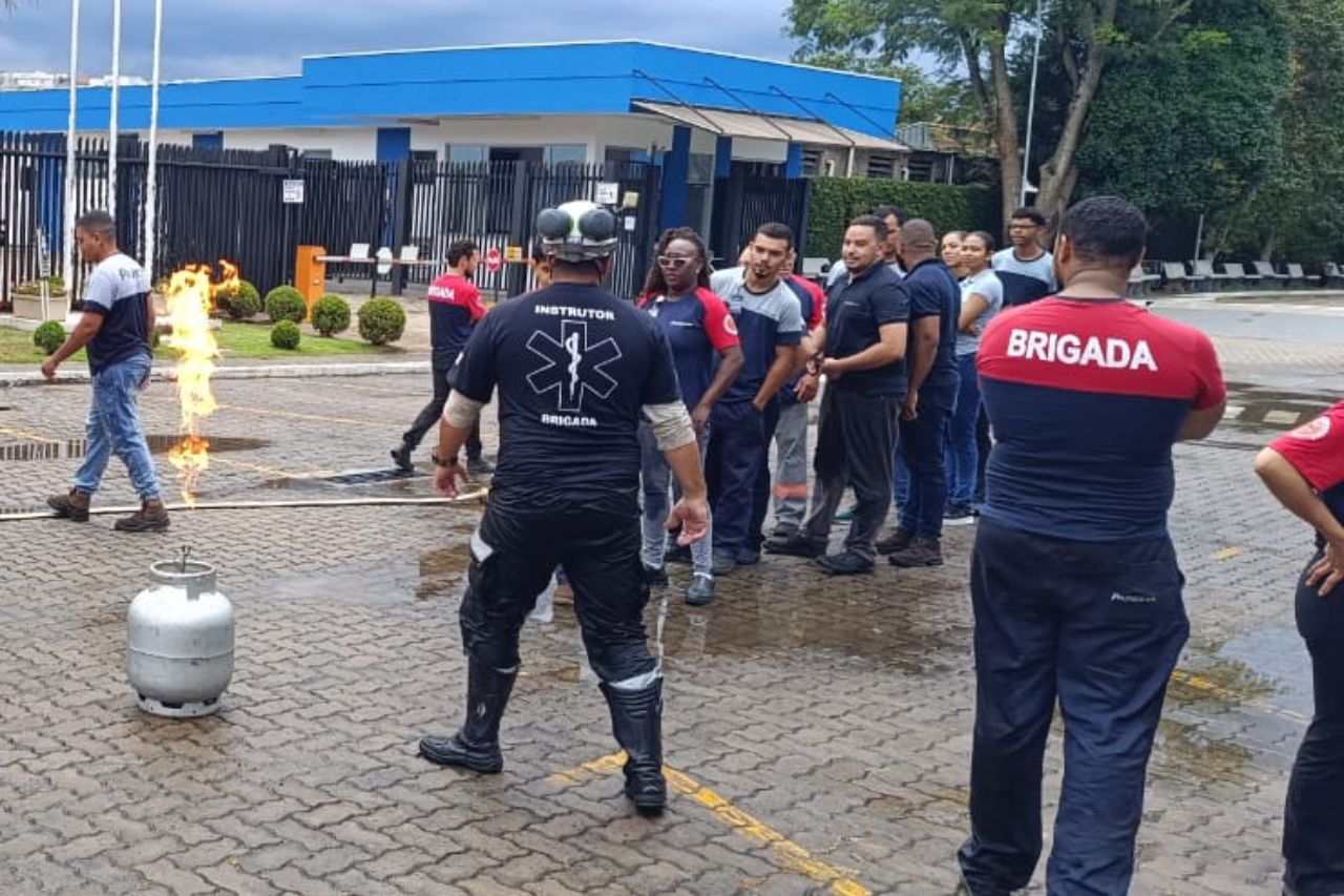 Bombeiros praticam o combate a um incêndio a gás ao ar livre, enquanto um grupo observa.