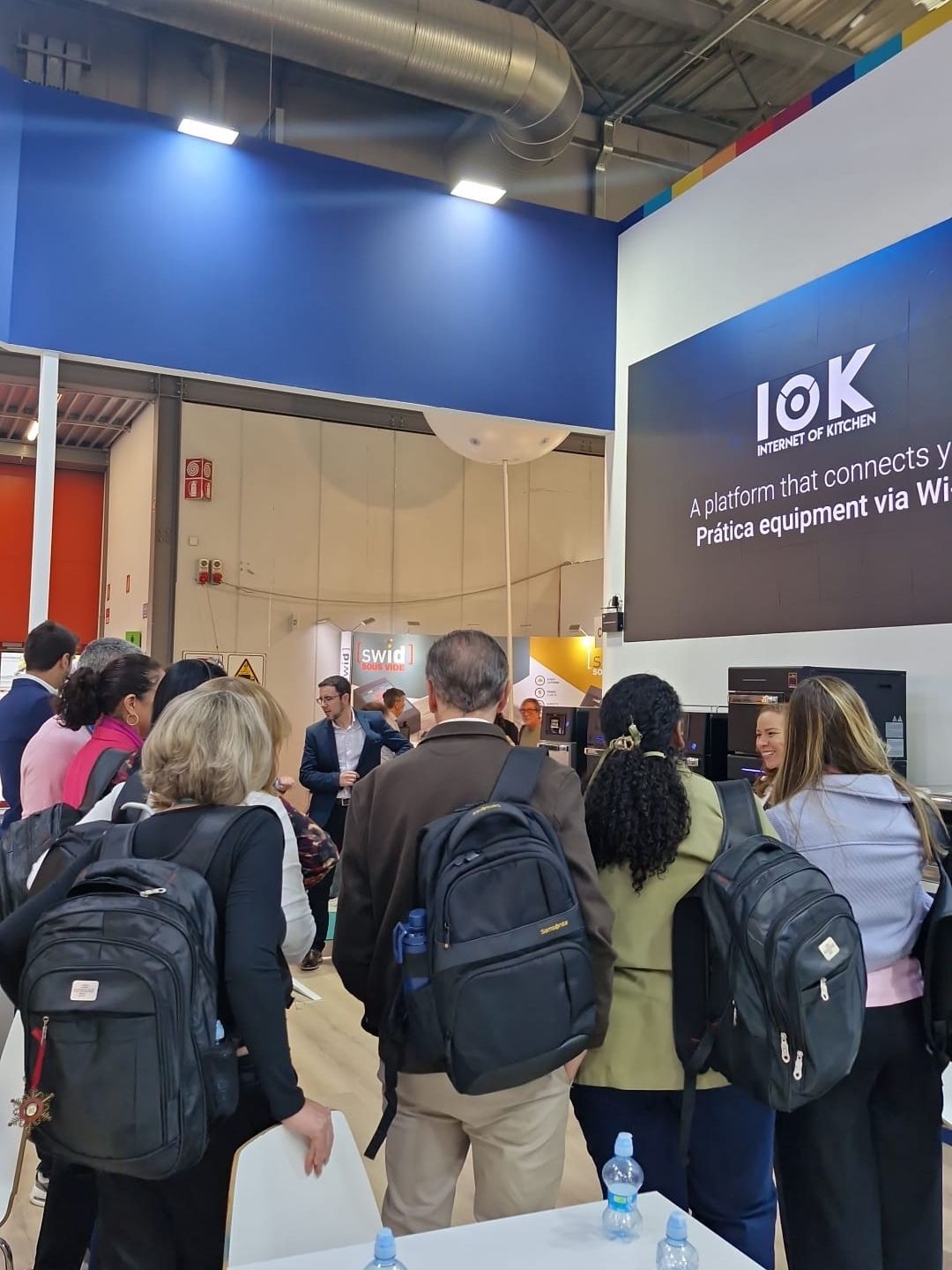 Pessoas em um estande de feira comercial, observando uma tela que diz 