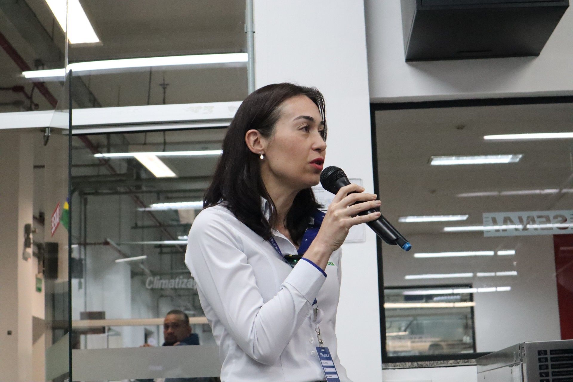Mulher de camisa branca falando ao microfone. Ambiente interno, com uma grande janela ao fundo.