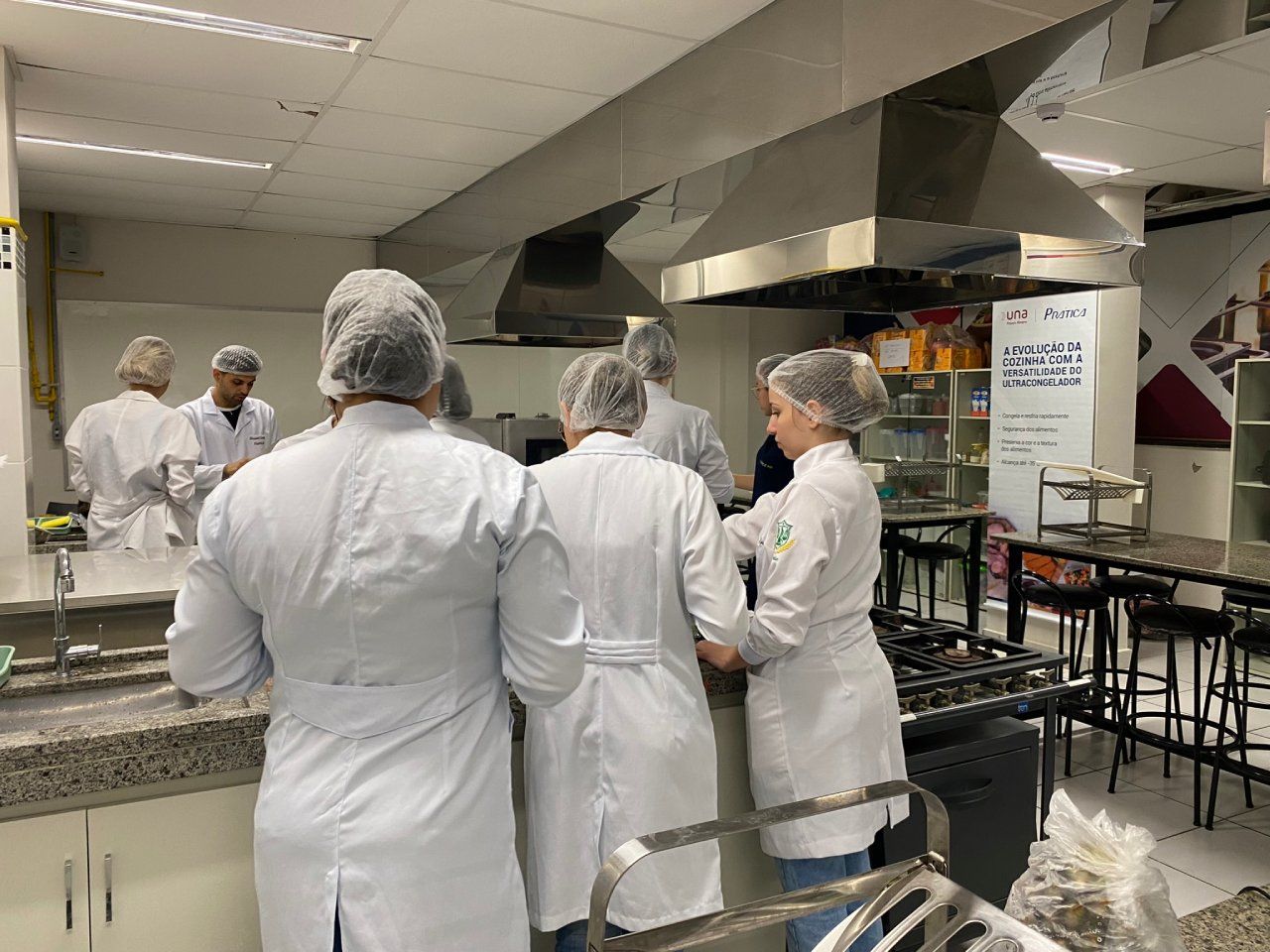 Um grupo de pessoas de jaleco branco e rede no cabelo estão em uma cozinha.