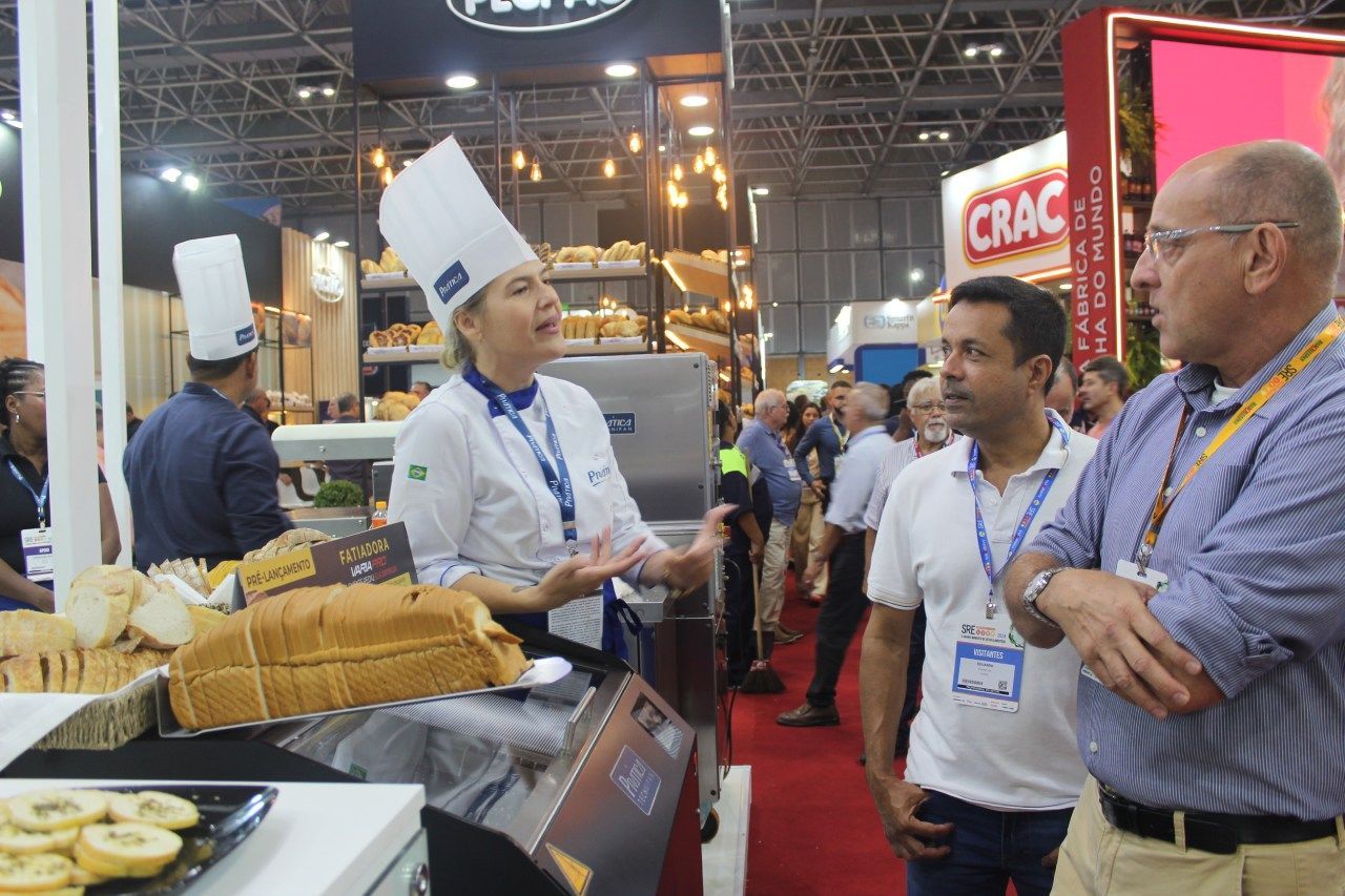 Uma mulher com chapéu de chef está conversando com dois homens em um show gastronômico.