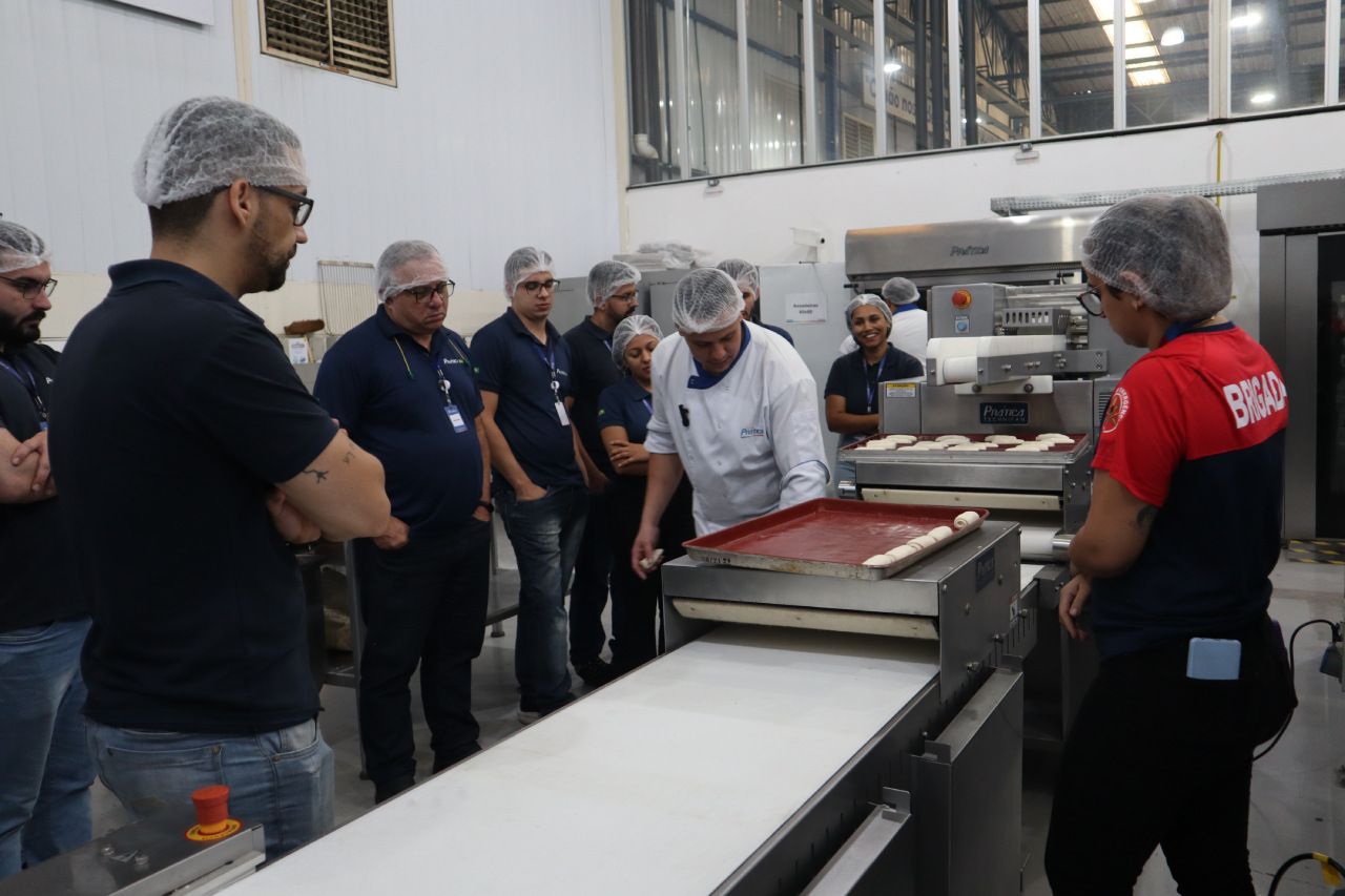 Um grupo de pessoas está em pé ao redor de uma esteira rolante em uma padaria.