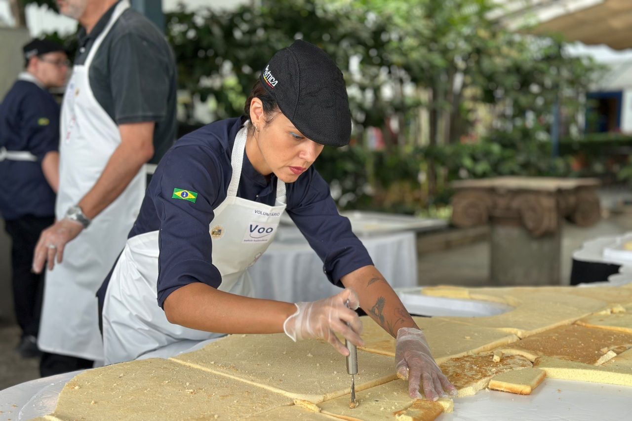 Mulher vestida com uniforme de chef cortando quadrados de bolo ao ar livre, usando touca e luvas. Outras pessoas aparecem ao fundo.