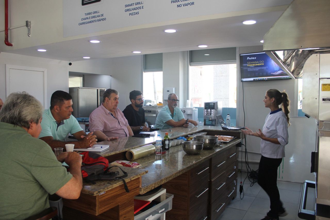 Um grupo de pessoas está sentado à mesa na cozinha enquanto uma mulher conversa com elas.