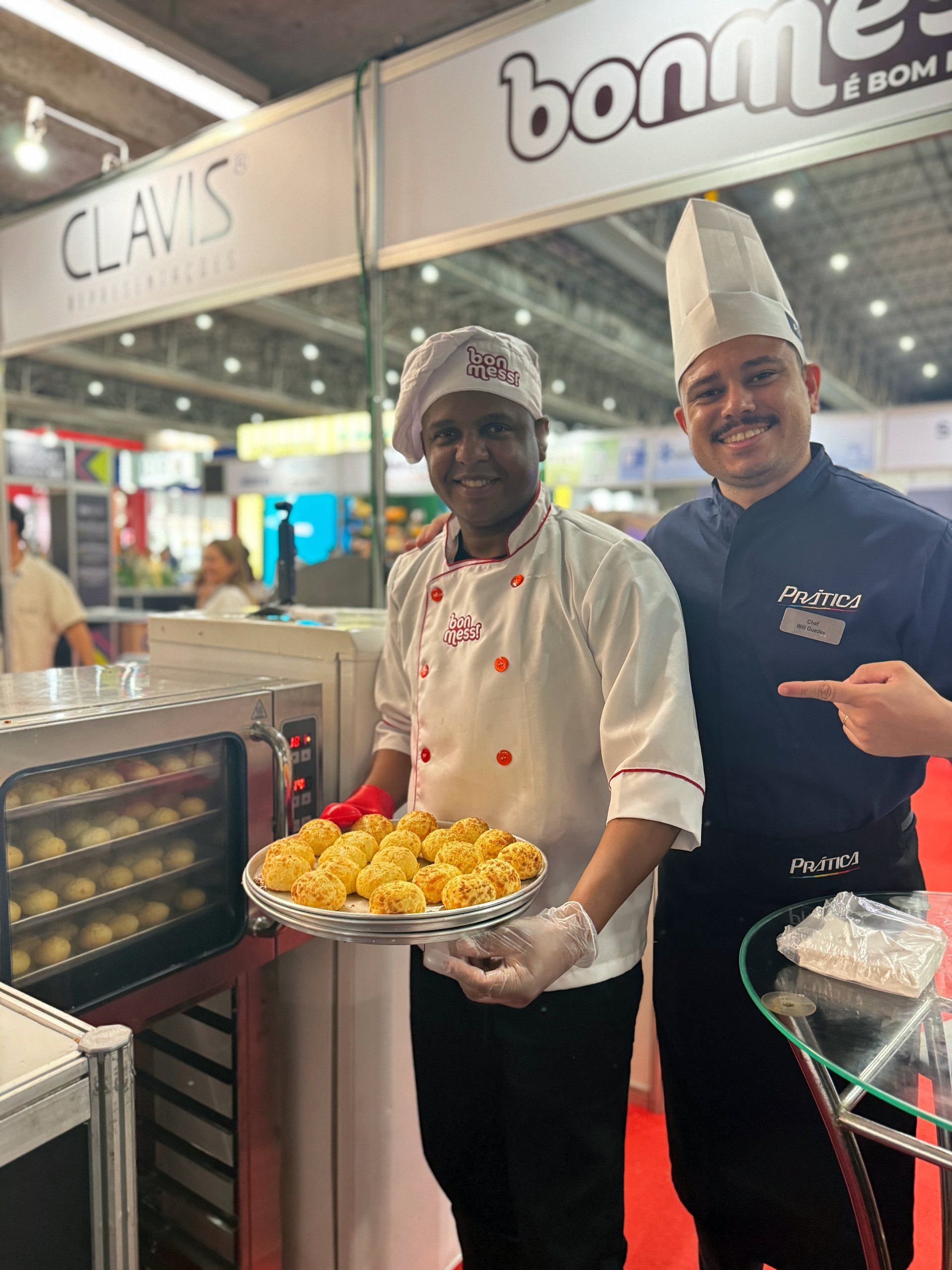 Dois chefs sorrindo enquanto seguram produtos assados ​​em um estande de feira comercial com um forno.