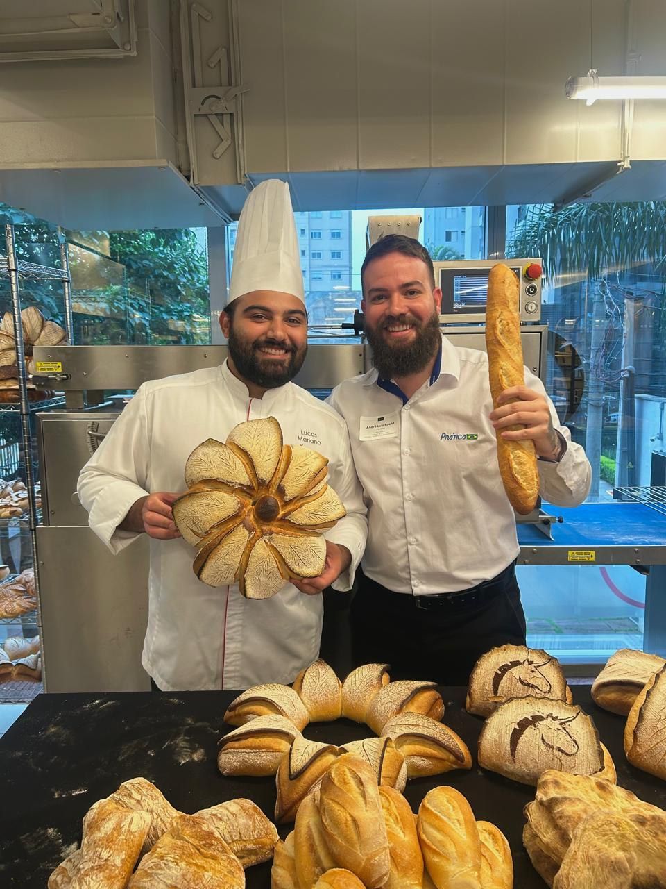 Dois homens estão de pé, um ao lado do outro, segurando pão em uma padaria.