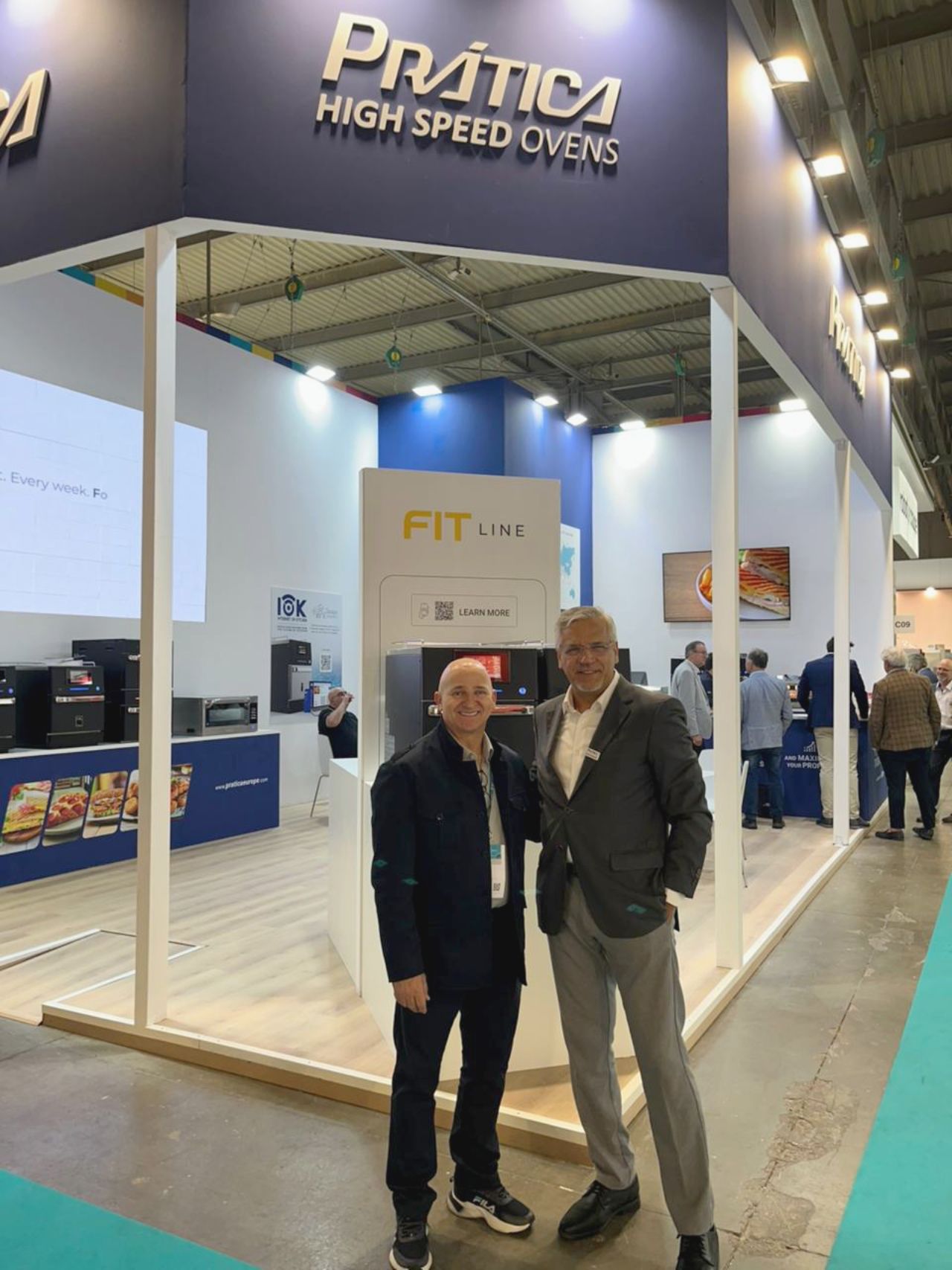 Dois homens estão em frente a um estande de uma feira comercial que diz 