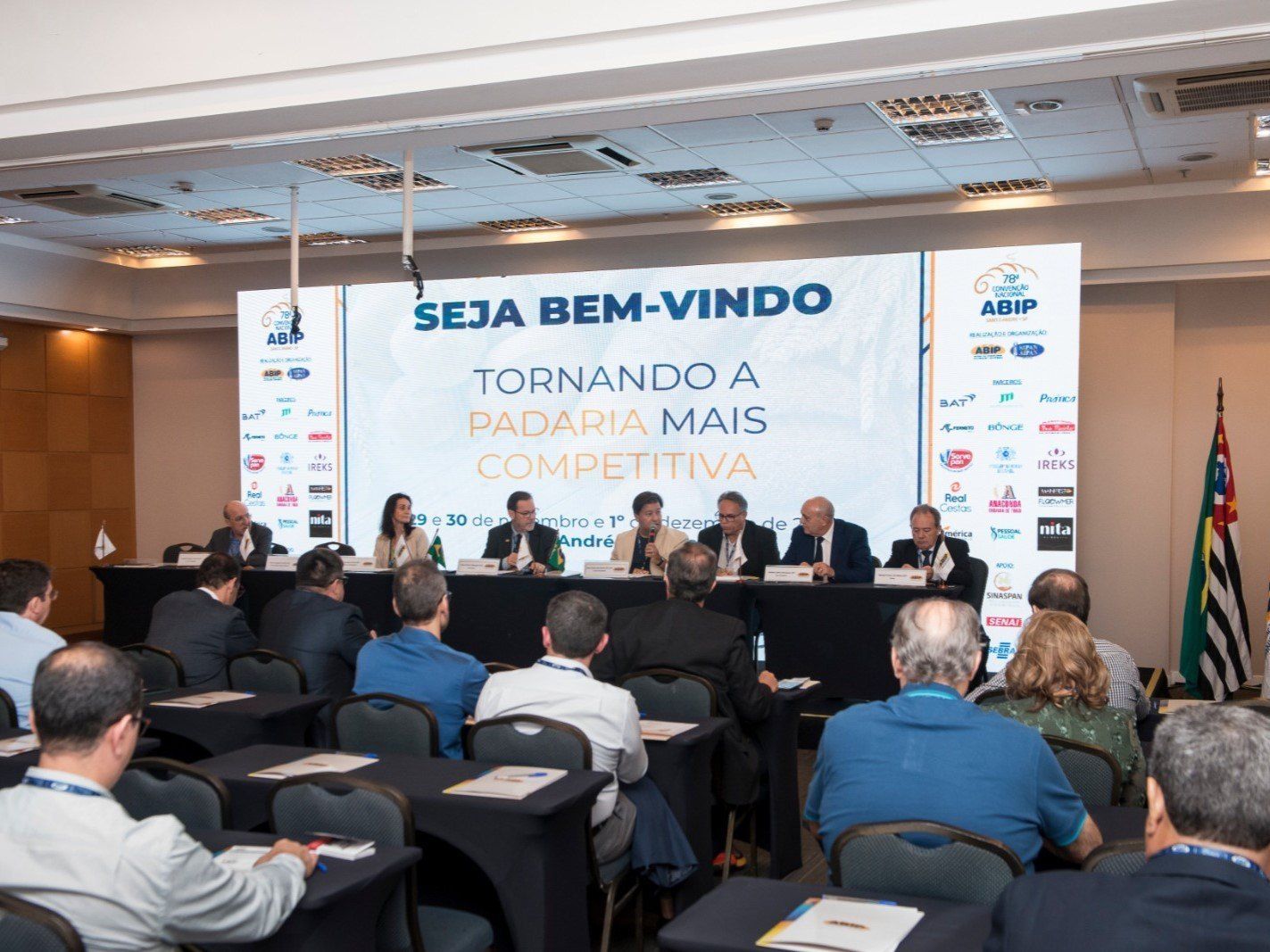 Um grupo de pessoas está sentado em mesas em frente a uma placa que diz seja bem-vindo