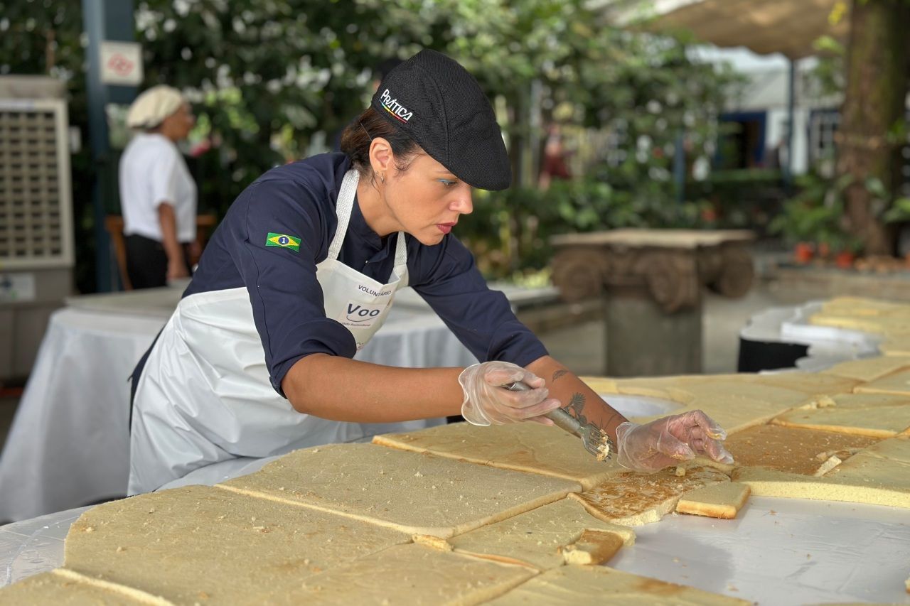 Chef uniformizado cortando quadrados de bolo em uma grande mesa, em um ambiente externo.