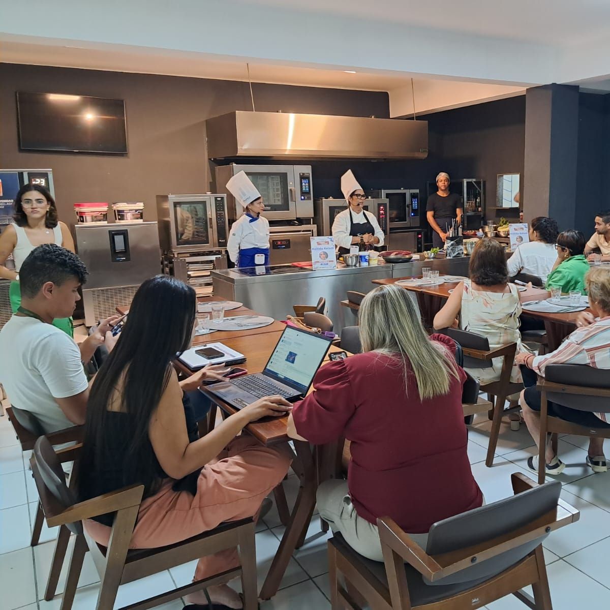 Um grupo de pessoas está sentado em mesas com laptops em uma cozinha.