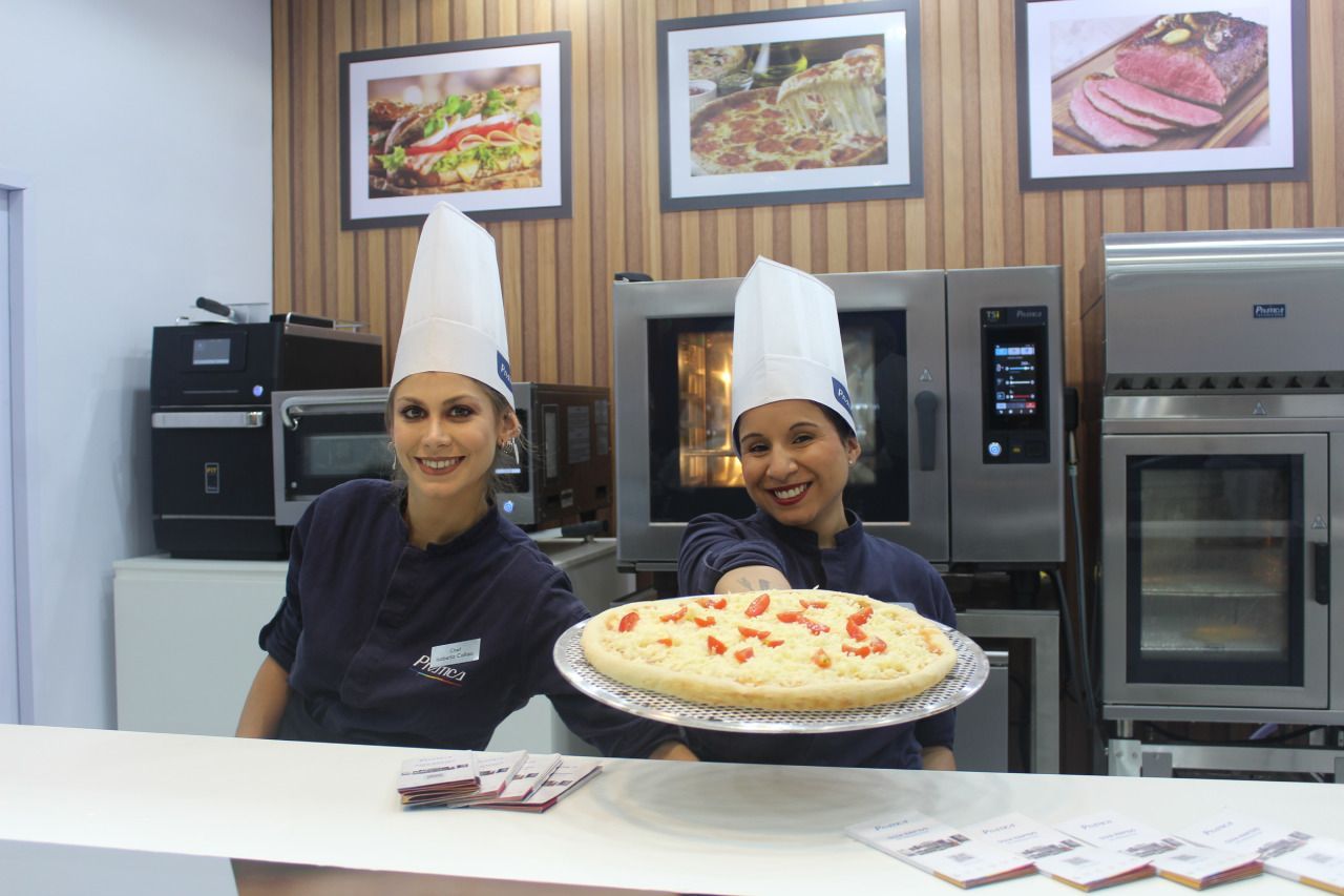 Duas mulheres com chapéus de chef segurando uma pizza em uma cozinha