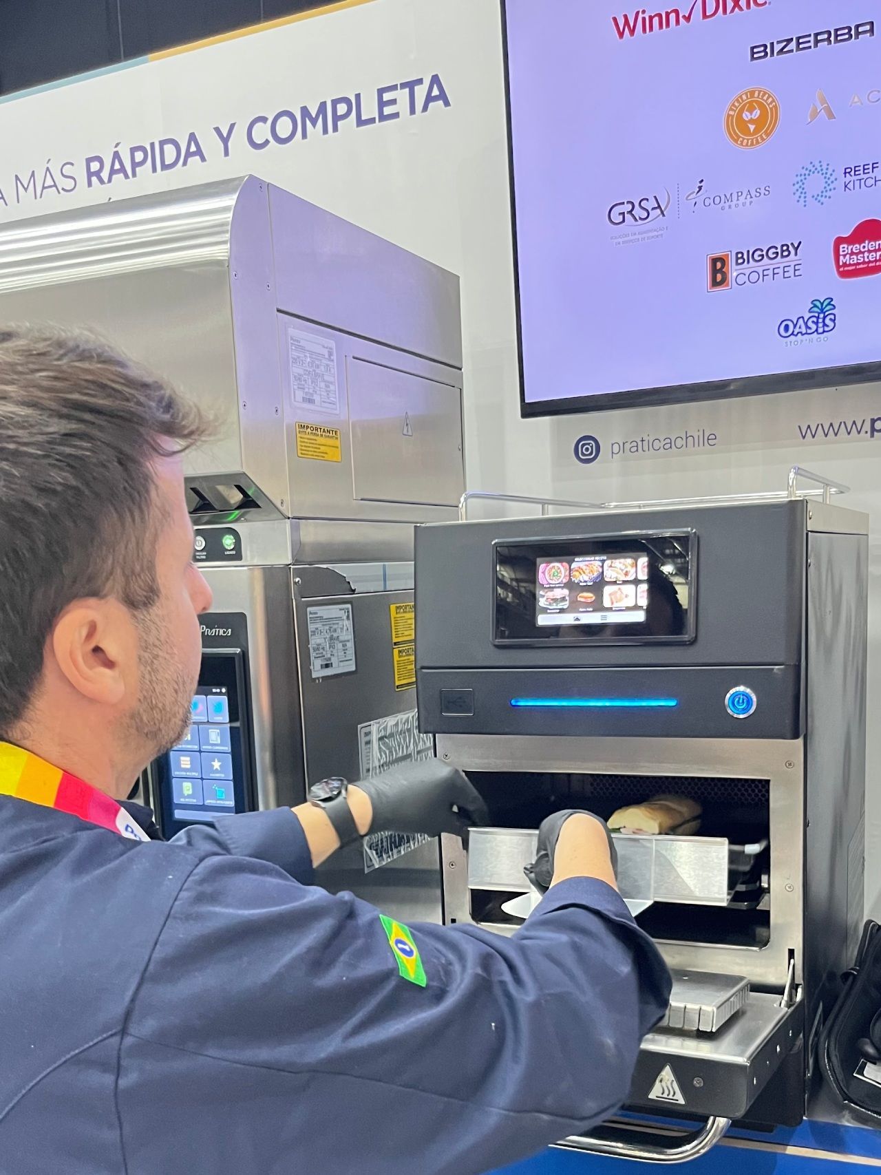 Homem com jaleco azul de chef colocando itens em um forno comercial em um evento, olhando para a exposição do forno.