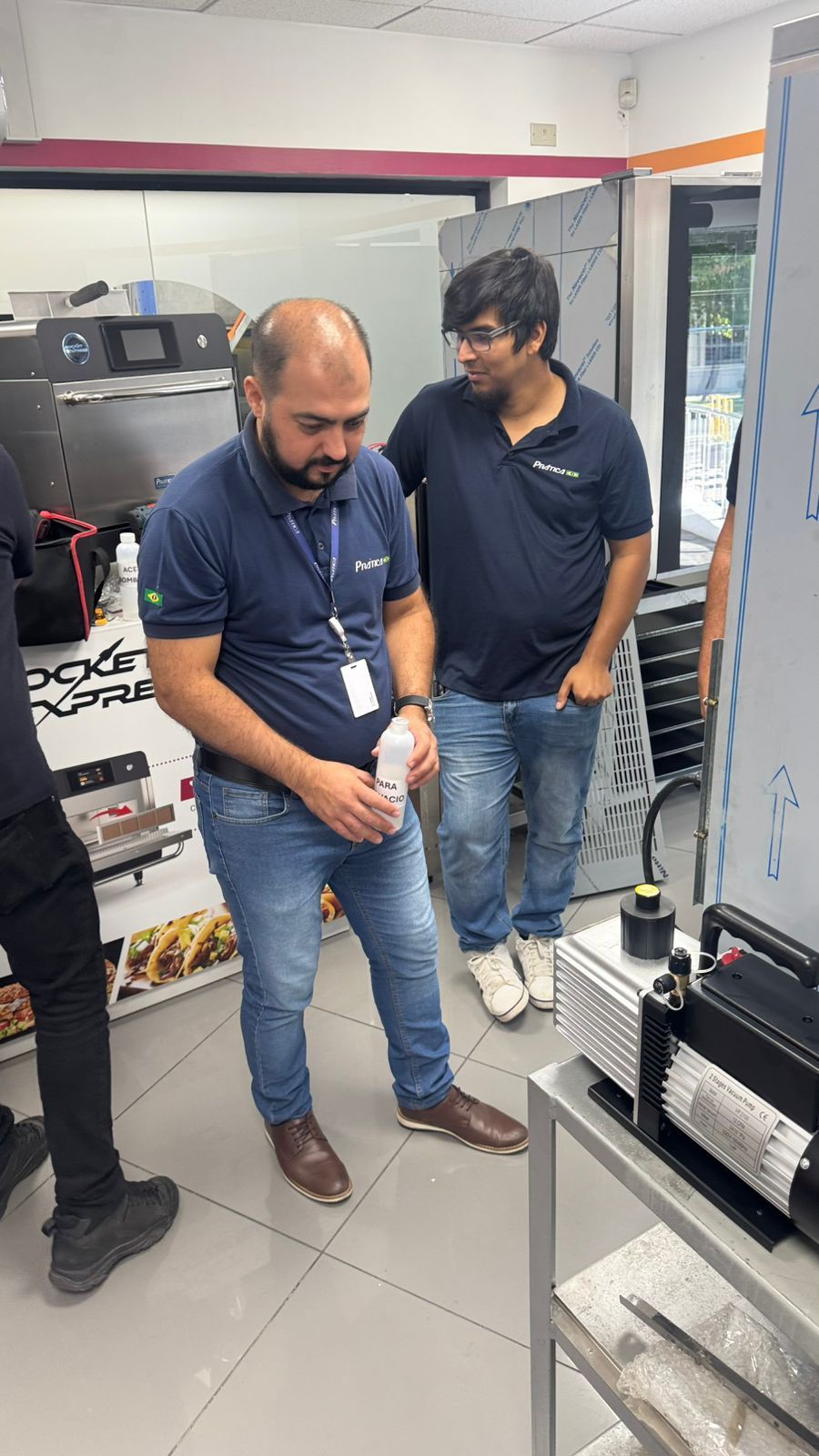 Dois homens em uma loja observando máquinas. Um deles segura uma garrafa; ambos estão vestidos casualmente.