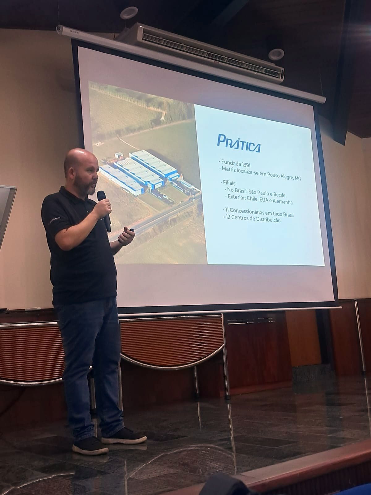 Homem apresentando para o público, apontando para um slide projetado com imagem de edifício e texto.