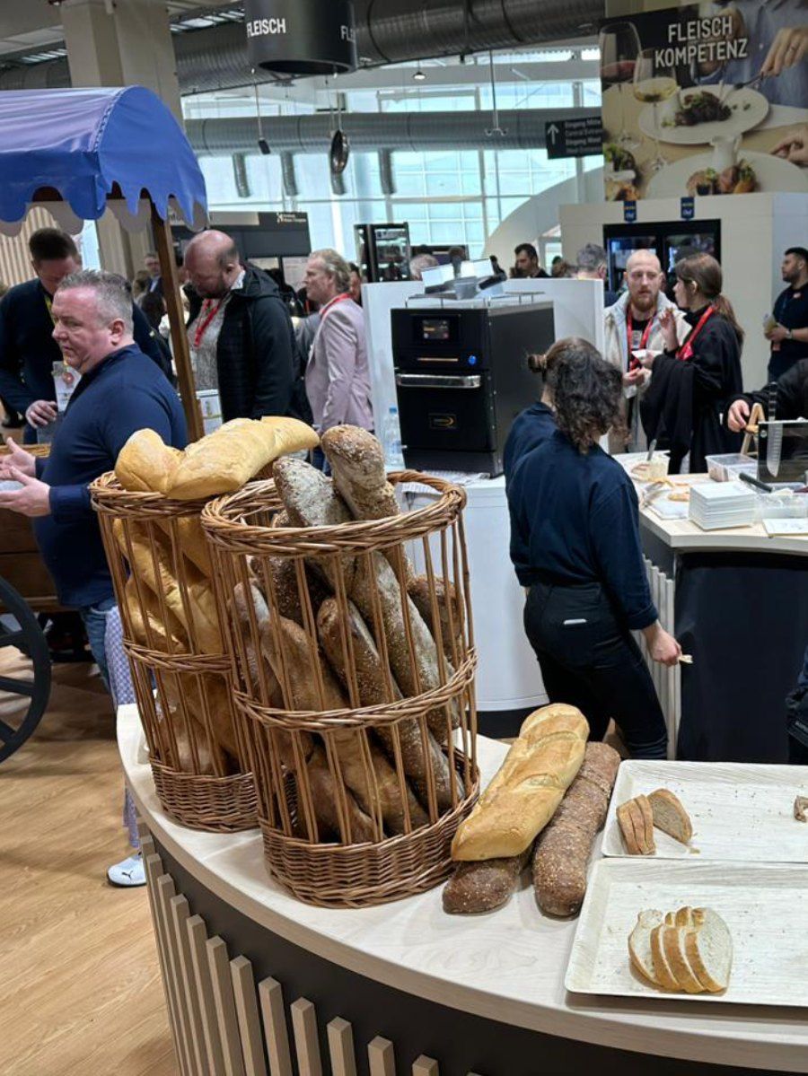 Um grupo de pessoas está em pé ao redor de uma mesa com cestas de pão sobre ela.