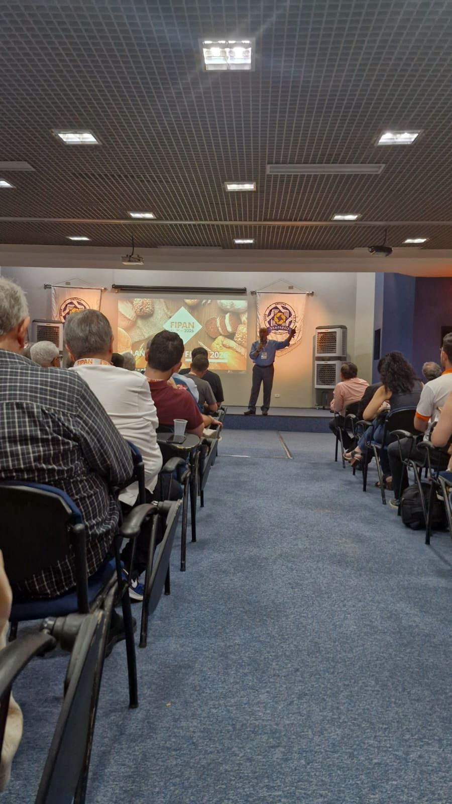 Um homem faz uma apresentação em uma conferência. Público sentado em fileiras, assistindo a uma apresentação em um palco com tela.