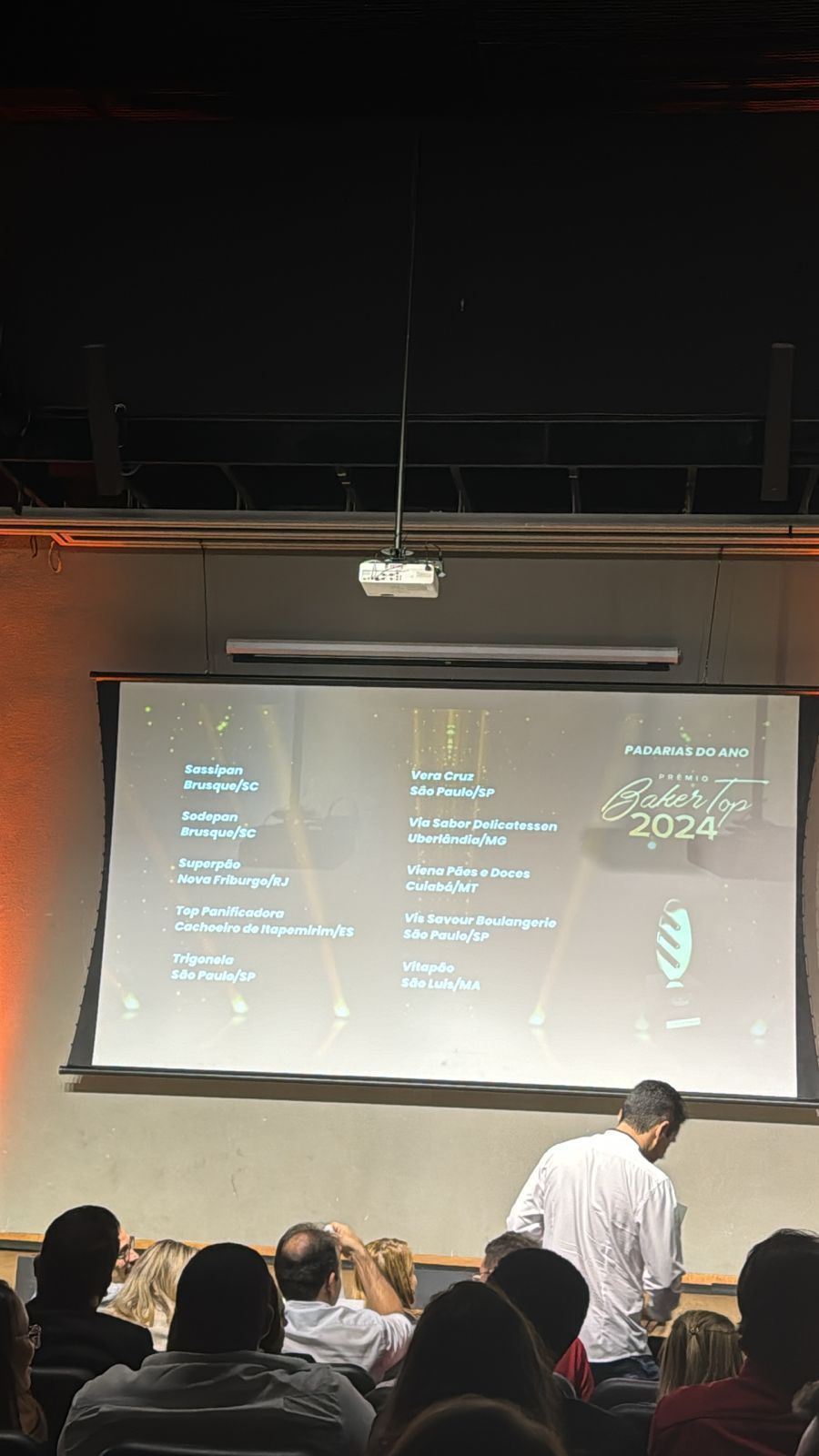 Um homem está fazendo uma apresentação para um grupo de pessoas em frente a uma tela grande.