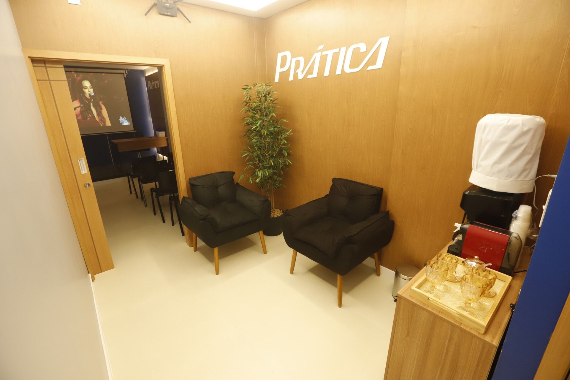 Área de estar com duas poltronas pretas, planta, bebedouro e porta para uma sala com cadeiras e biombo.