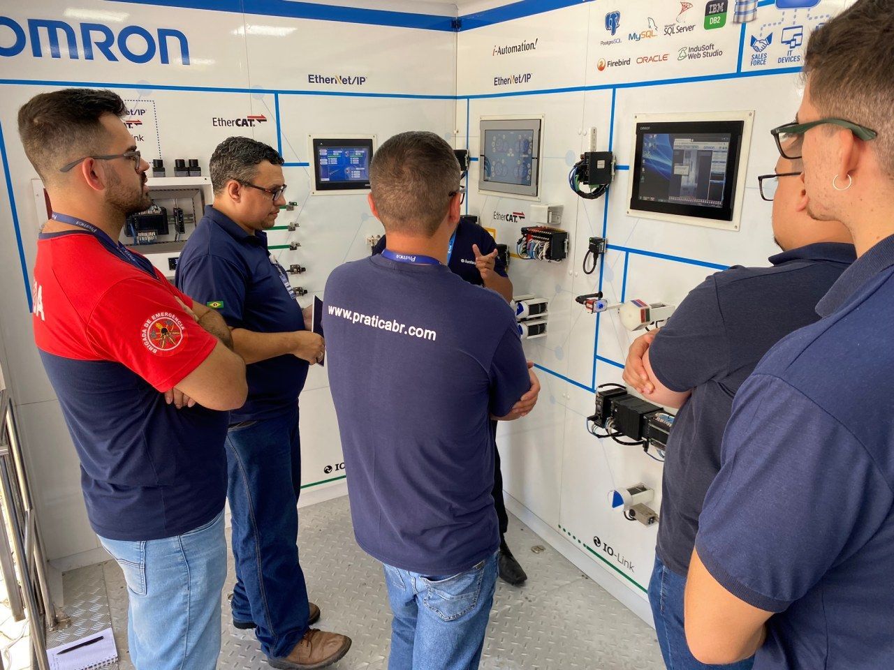 Um grupo de homens está parado em frente a uma parede onde está escrito Omron.