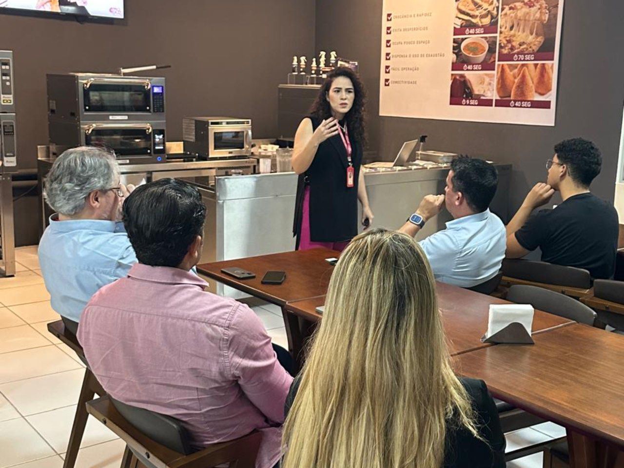 Uma mulher está fazendo uma apresentação para um grupo de pessoas sentadas em mesas em um restaurante.