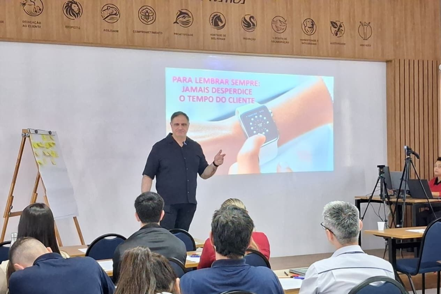 Um homem faz uma apresentação para um grupo. Ele aponta para a projeção de um relógio. As pessoas sentam-se às mesas.