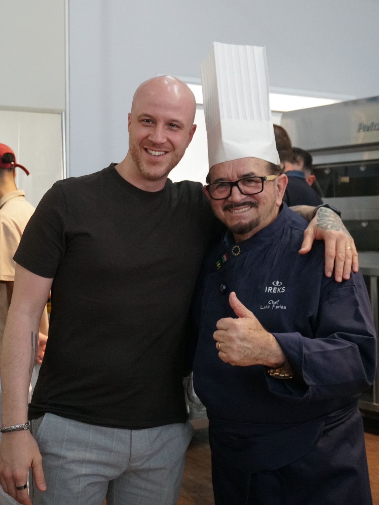 Dois homens, um sorrindo com a cabeça raspada, o outro, um chef com o polegar para cima. Eles posam em uma cozinha.