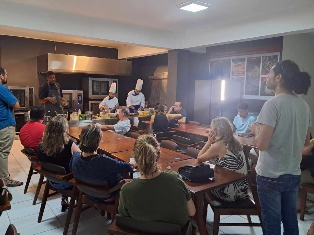 Um grupo de pessoas está sentado em mesas em uma sala assistindo a uma aula de culinária.