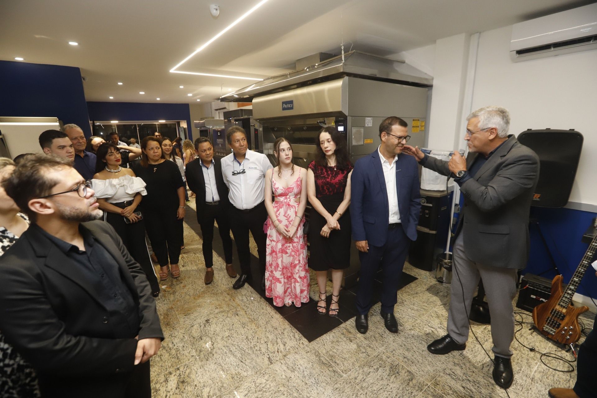 Um grupo de pessoas em um evento, com um homem falando. Em um ambiente interno, com um forno ao fundo.