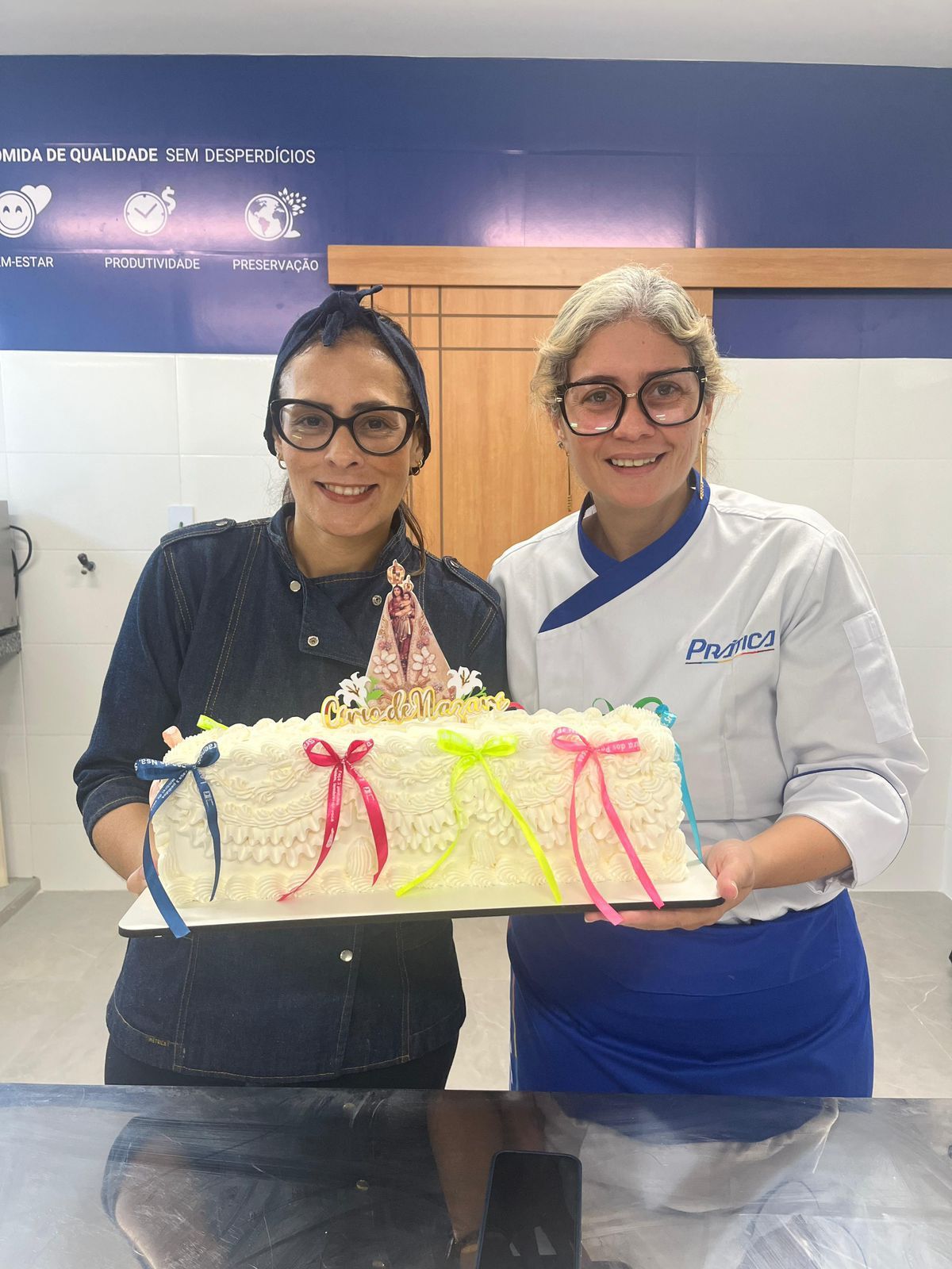 Duas mulheres sorrindo, segurando um bolo decorado em uma cozinha.