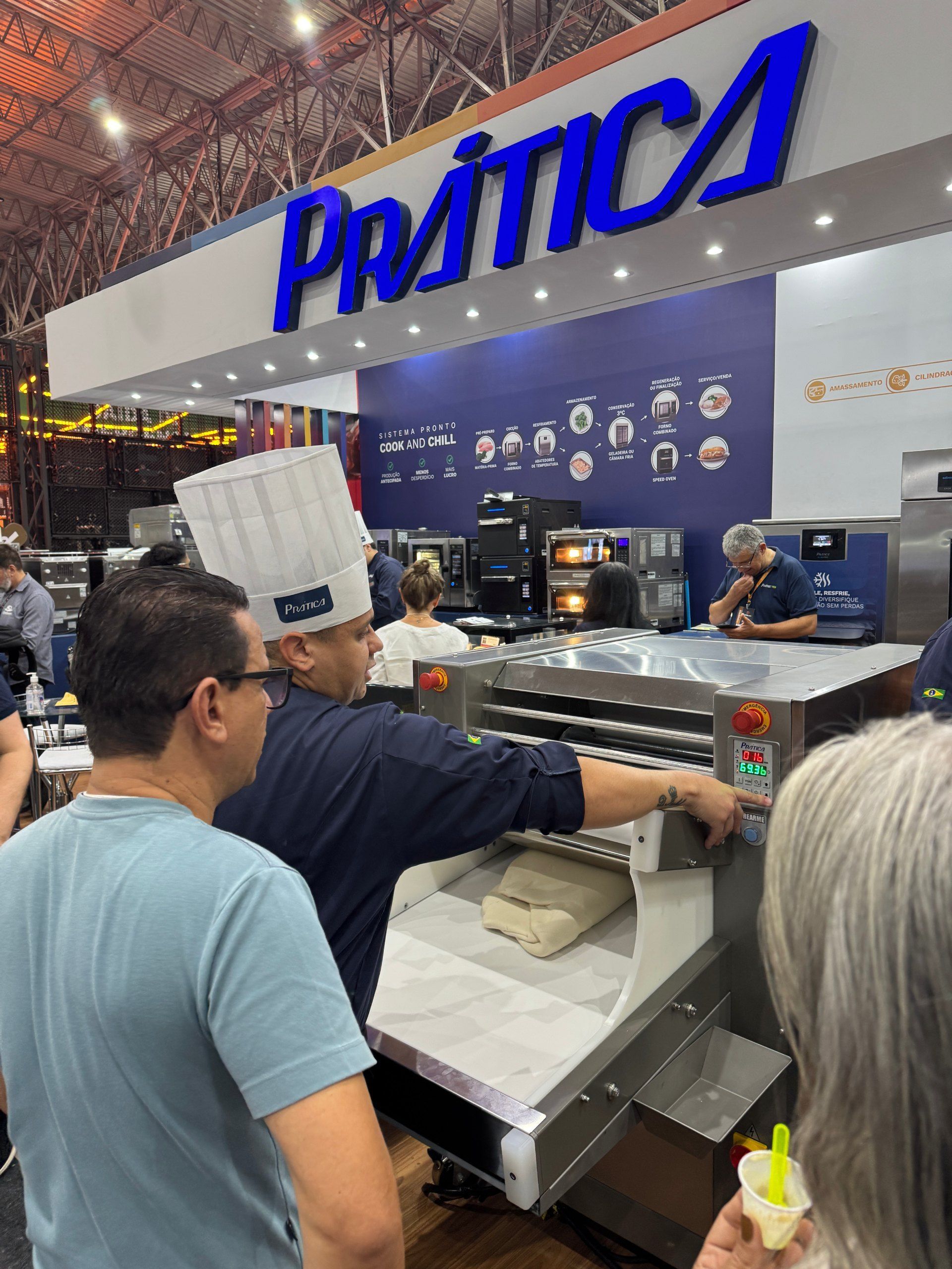 Um chef demonstra o funcionamento de uma máquina de processamento de alimentos em um estande de feira comercial, sob o olhar atento de espectadores.