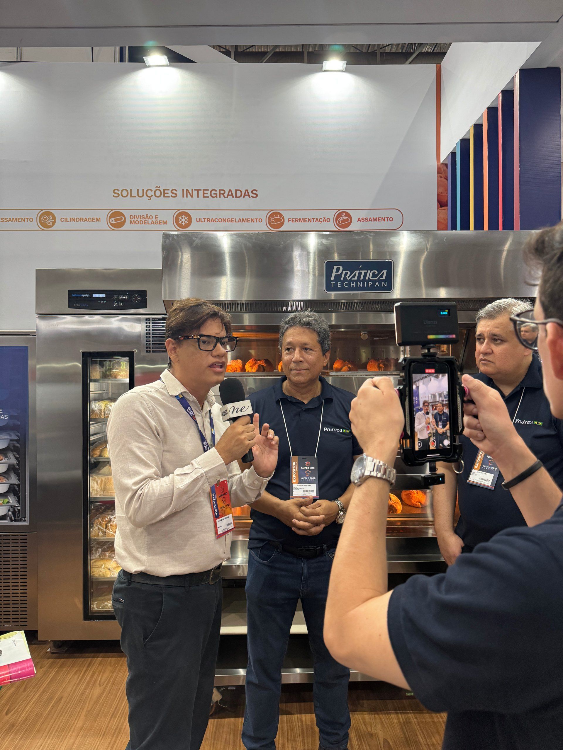 Um homem com um microfone entrevista dois homens em uma exposição de equipamentos de cozinha industrial.