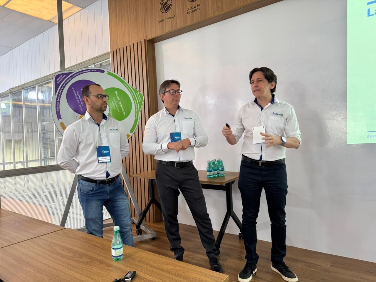 Três homens de camisa branca em uma apresentação, um deles falando com um microfone, em um palco com um logotipo.