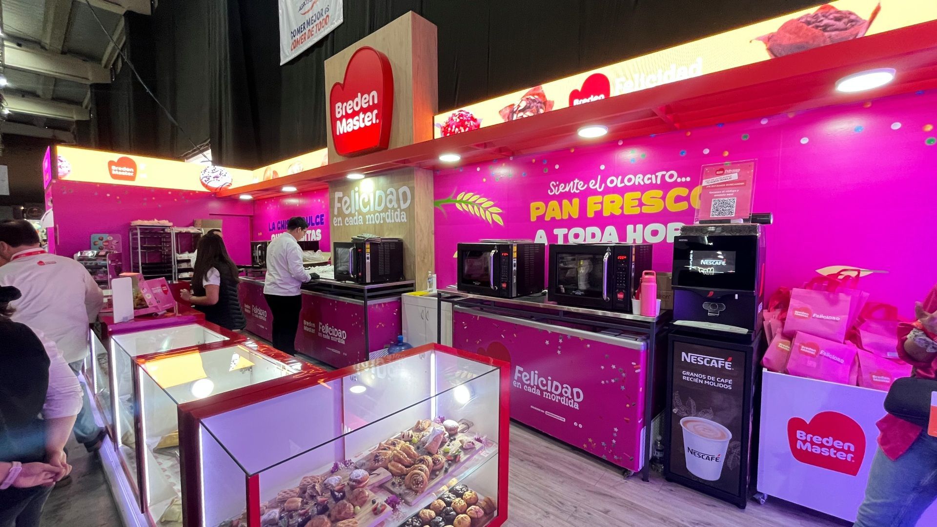 Um estande rosa de uma padaria em uma feira. Produtos assados em vitrines, fornos e funcionários presentes.