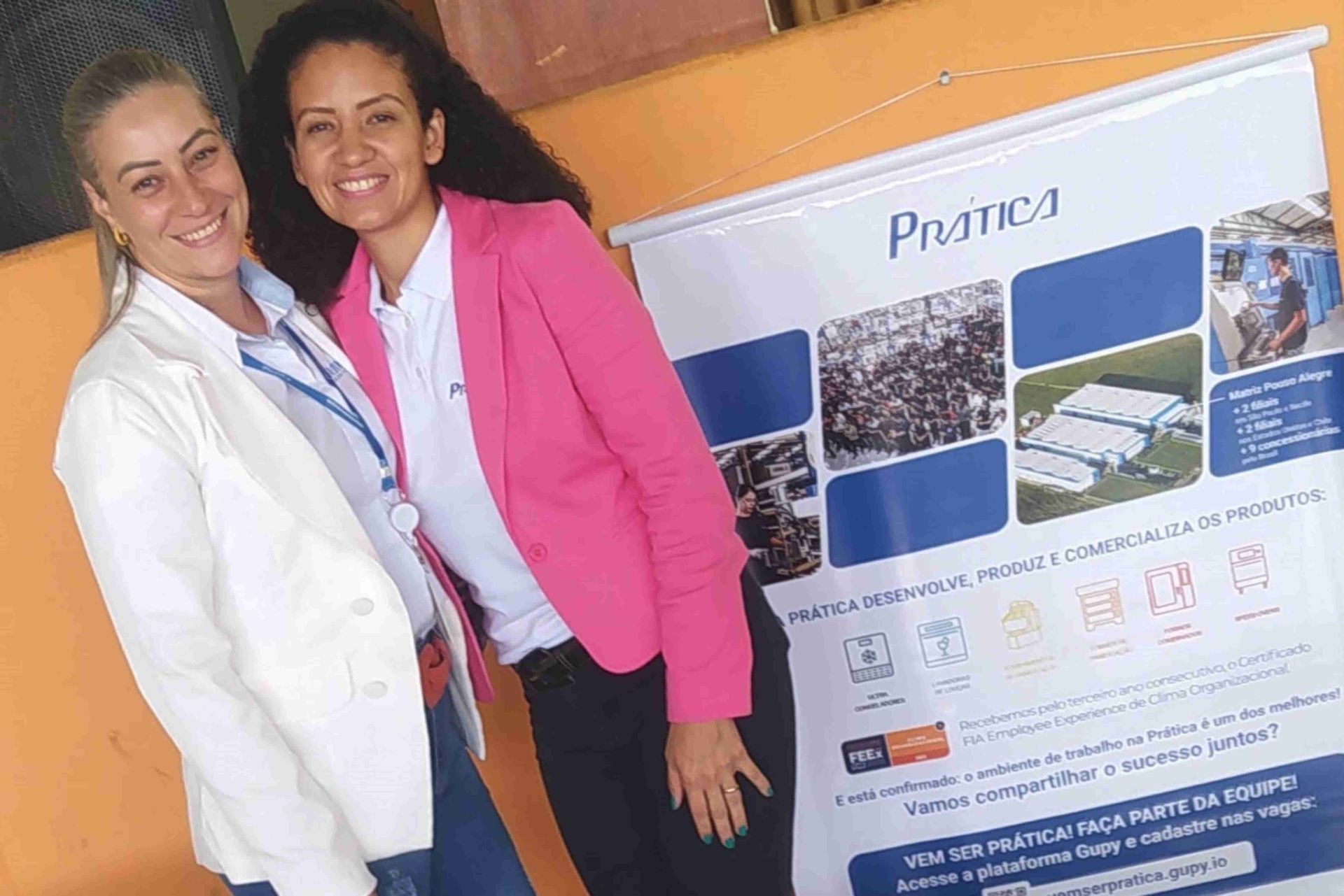 Duas mulheres estão de pé, uma ao lado da outra, em frente a uma faixa.