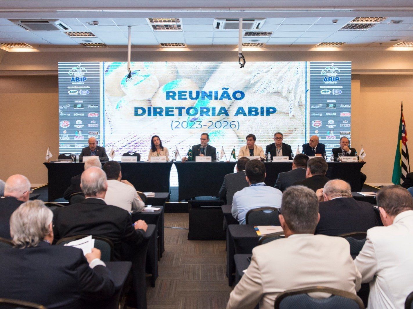 Um grupo de pessoas está sentado em frente a uma tela grande que diz reunião diretoria abip