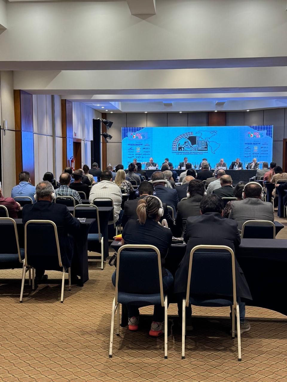 Um grande grupo de pessoas está sentado em mesas em uma sala de conferências.