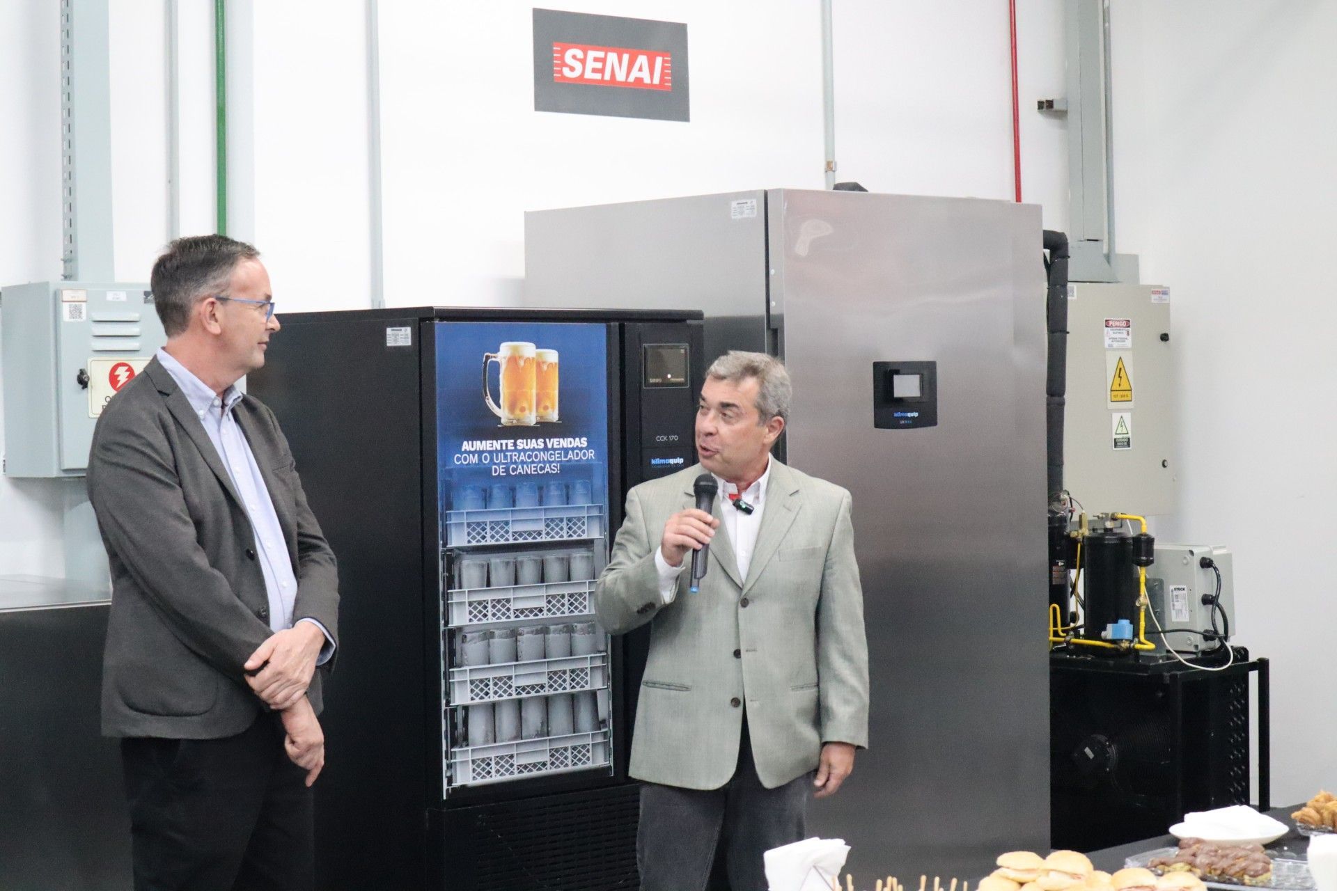 Dois homens ao lado de uma máquina de venda automática de cerveja, um deles falando ao microfone, em uma sala com a placa SENAI.