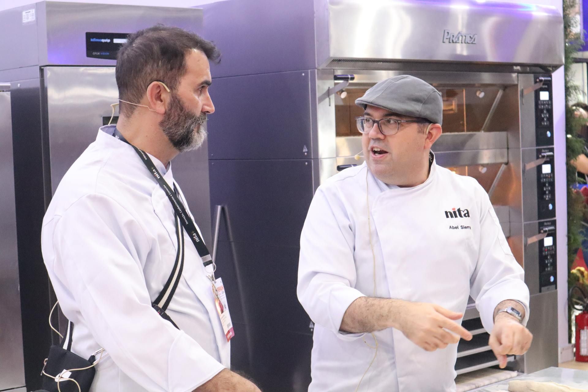 Dois homens estão parados um ao lado do outro em uma cozinha conversando entre si.