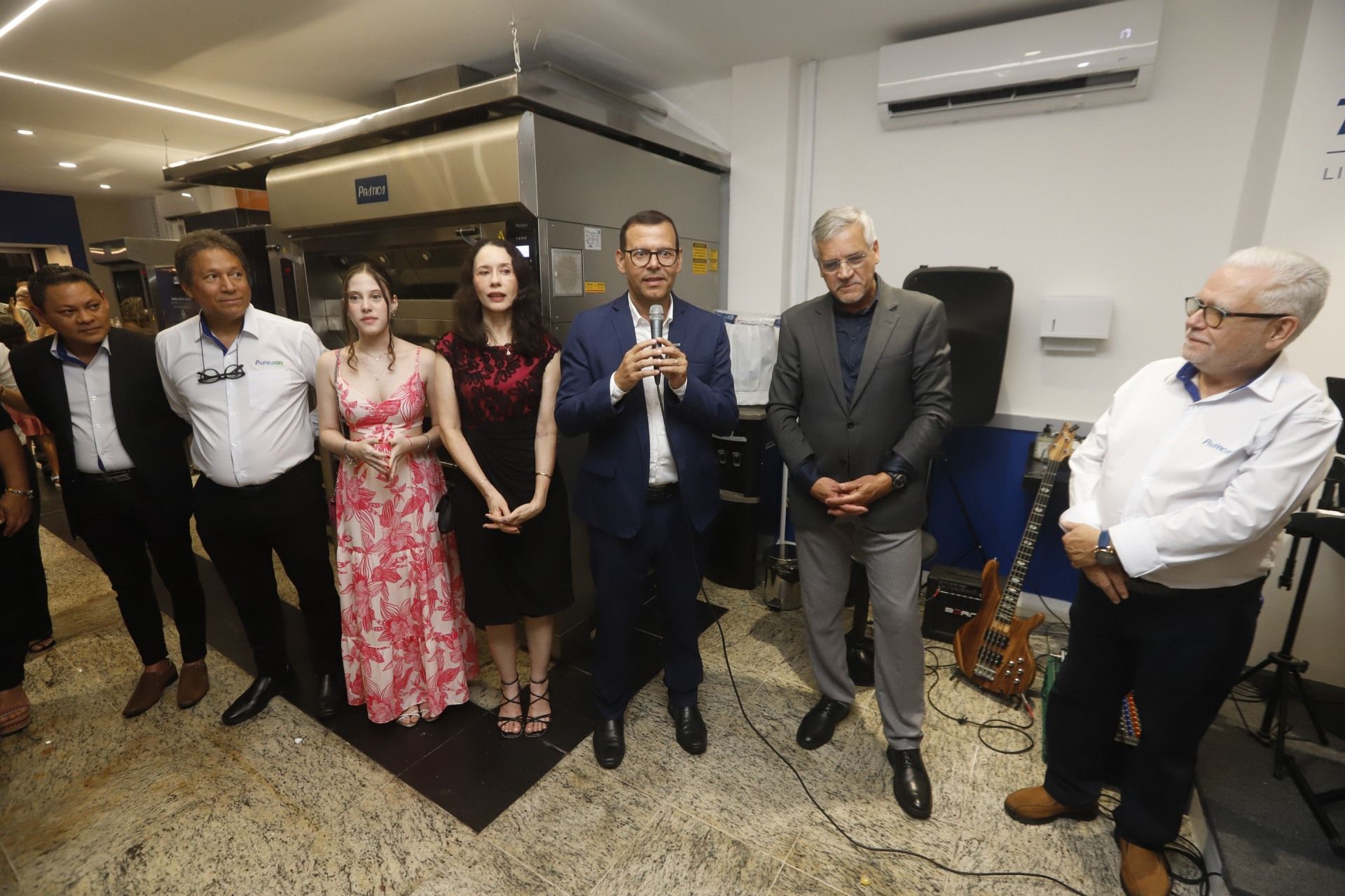 Grupo de pessoas em um evento, homem falando em um microfone, perto de um grande forno.