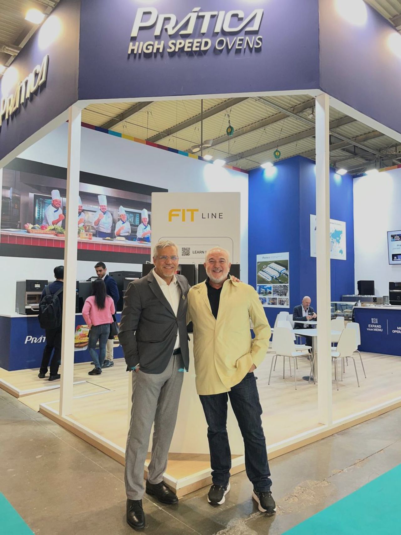 Dois homens posam em frente ao estande de fornos de alta velocidade da Pratica em uma feira comercial.