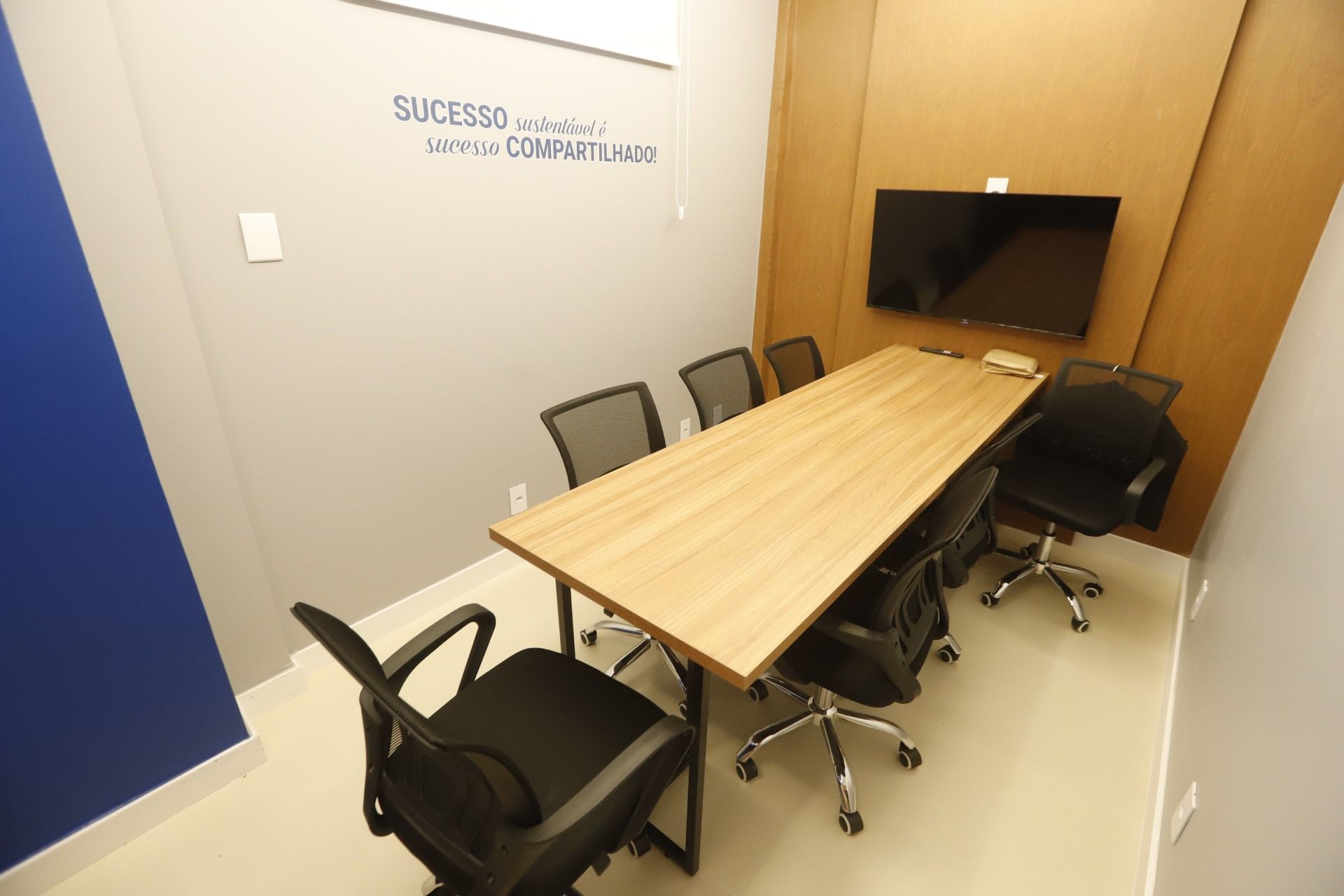 Sala de conferências com mesa longa, cadeiras, TV e texto motivacional na parede; tons de cinza, azul e madeira.