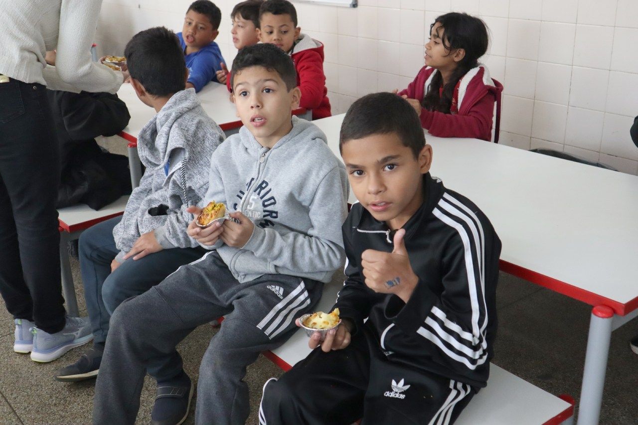 Um grupo de crianças está sentado em mesas em uma cafeteria e um menino está dando um sinal de positivo