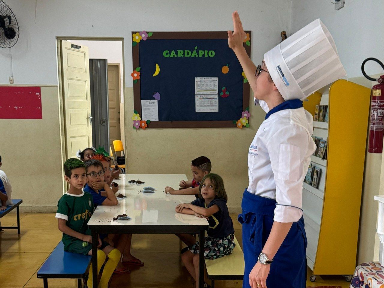 Uma mulher com um chapéu de chef está parada na frente de um grupo de crianças sentadas em mesas.
