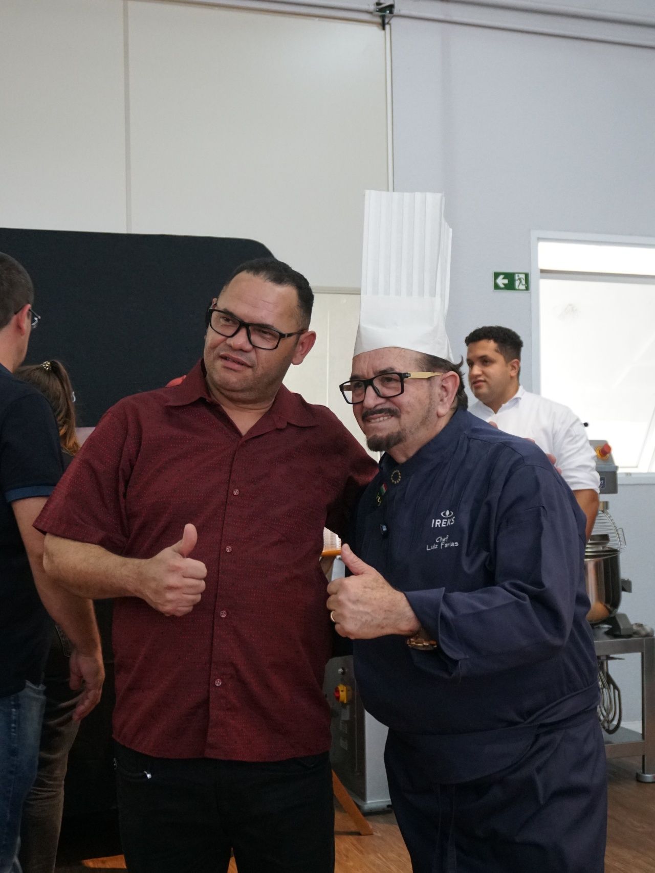 Dois homens sorrindo, fazendo sinal de positivo; um com uniforme de chef e chapéu, o outro com camisa vermelha. Dentro de uma cozinha.