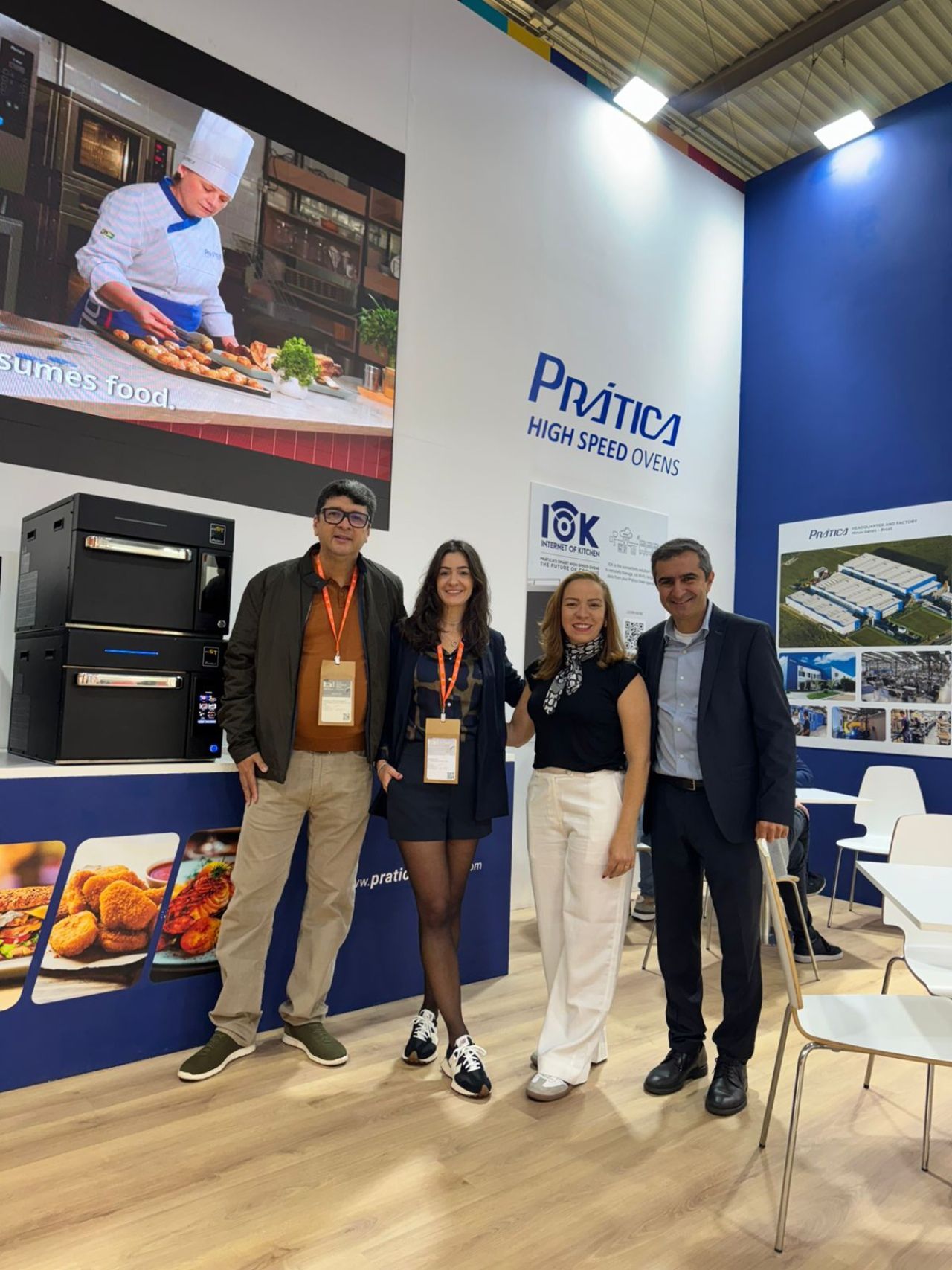 Quatro pessoas posam em um estande de feira comercial com equipamentos de cozinha e um logotipo.