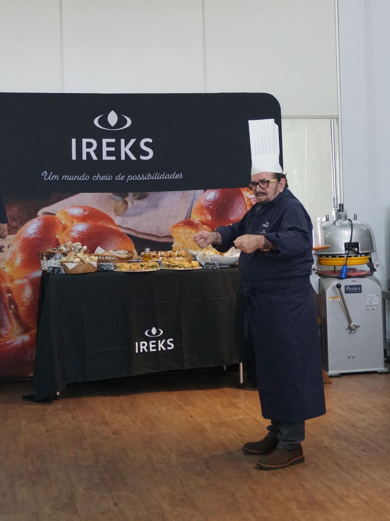 Chef de avental azul-marinho e touca gesticula em direção a uma exposição de produtos assados ​​em um evento da marca IREKS.