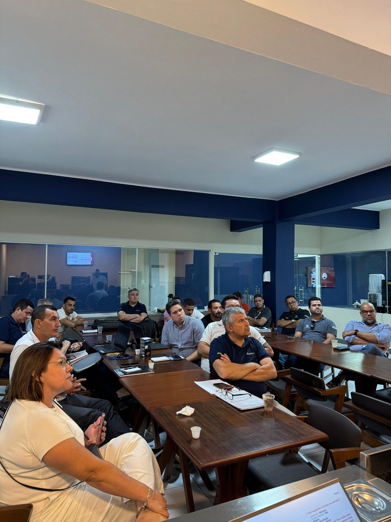 Pessoas sentadas ao redor de mesas em uma sala, assistindo a uma apresentação. A sala tem paredes azuis e uma janela com vista para a cidade.