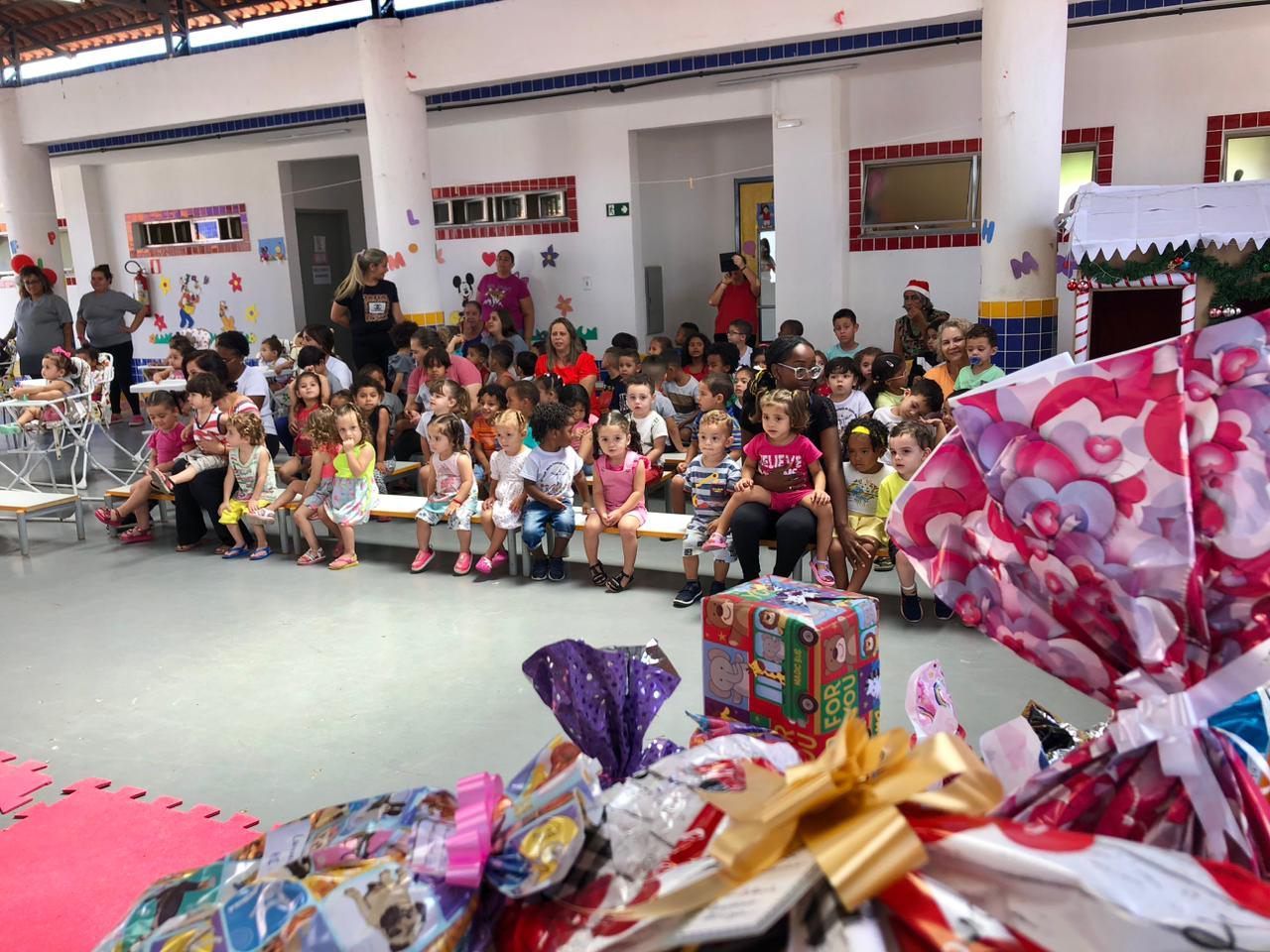 Um grupo de crianças está sentado em um banco em uma sala cercado por presentes.